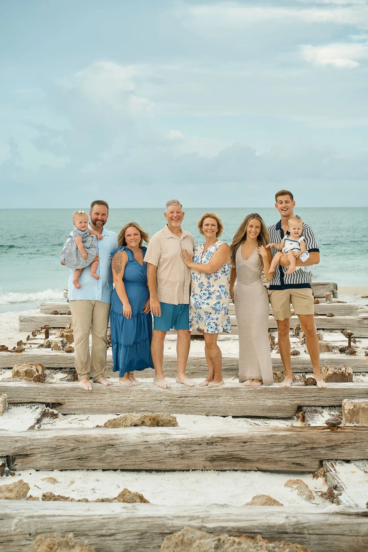 A family of eight standing barefoot in a row on the weathered timbers of an old pier by the sea: grandparents in the middle, four younger adults beside them and two babies held by two younger men, a pale blue sky with white clouds above
