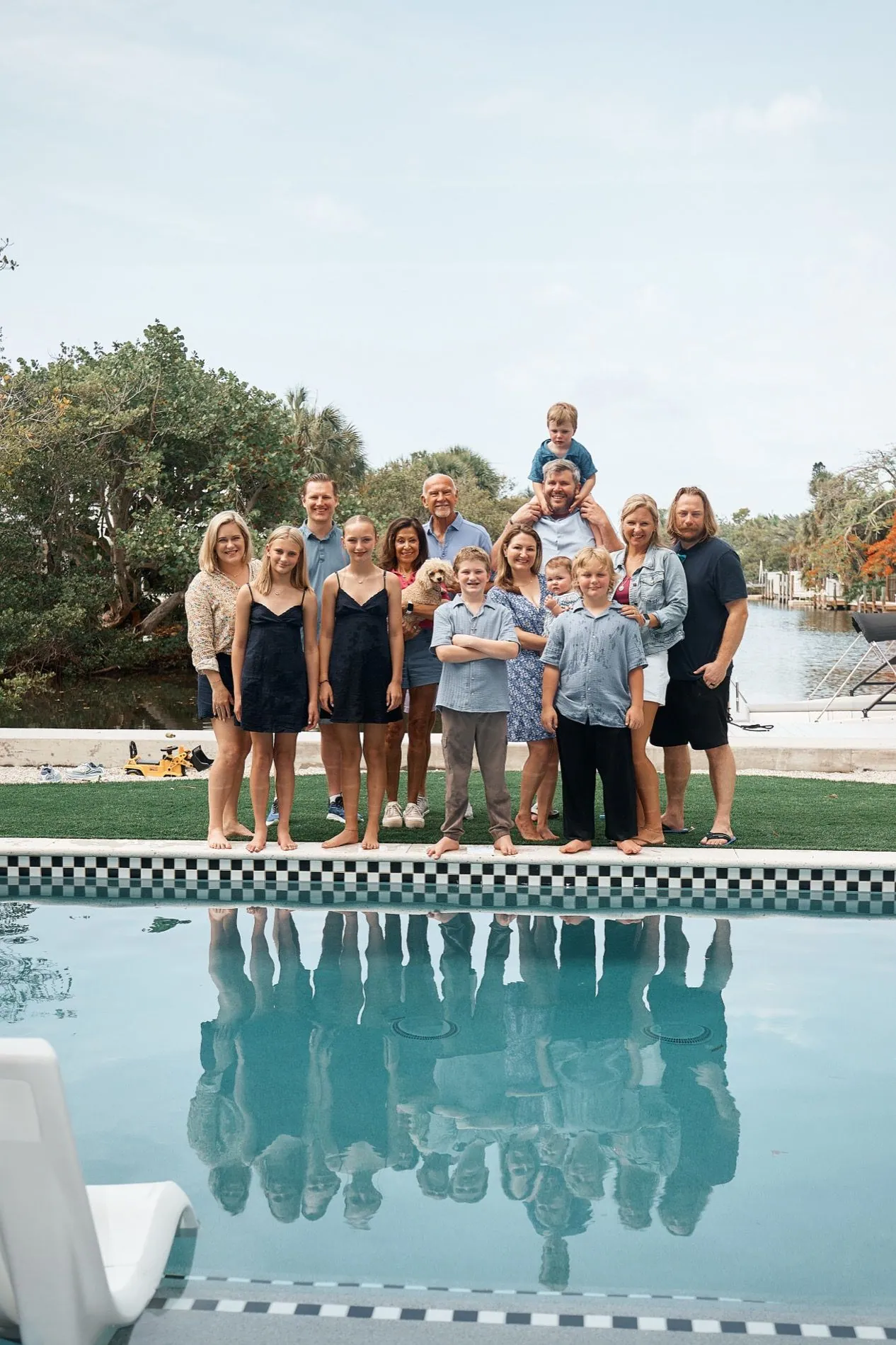 A big family standing in a row at the edge of a pool at a rental house, a small boy on a man's shoulders at the back, all of them reflected in the water