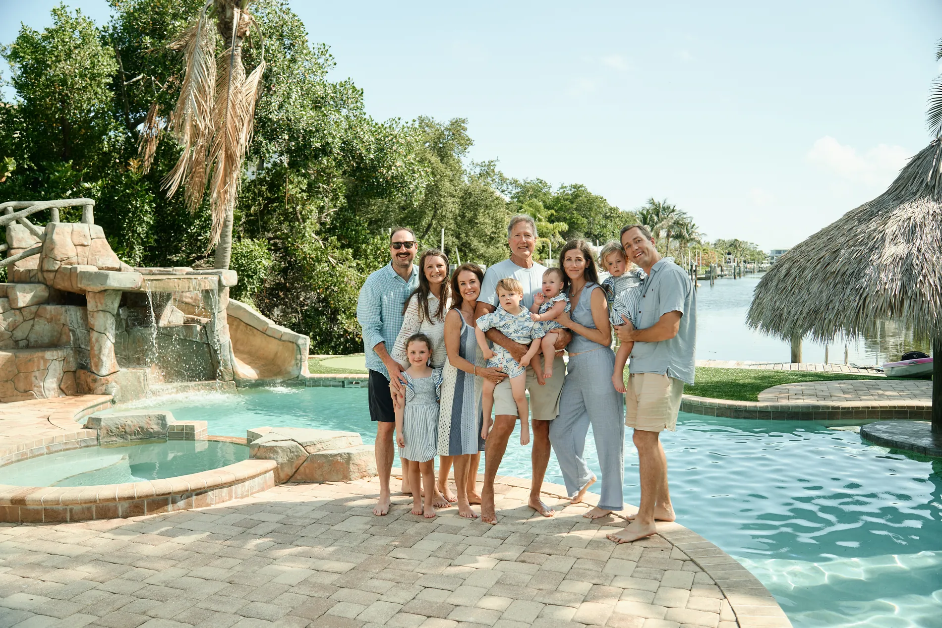 Three generations of one big family, dressed in light blue and white, standing together beside the pool of a rental house, a rock waterfall with a slide on one side and a thatched tiki hut on the other