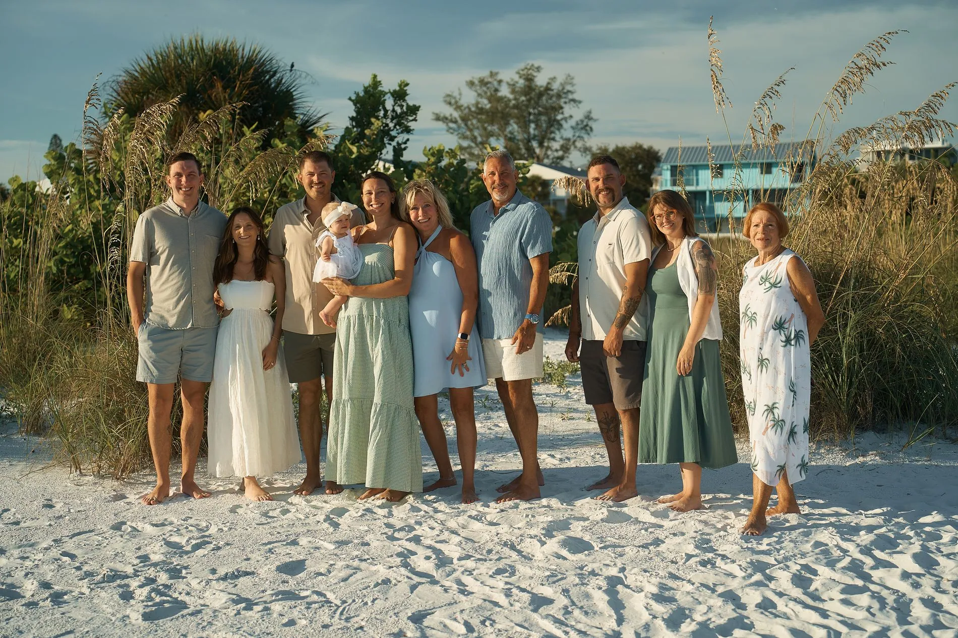 Eleven adults and a baby of one extended family standing in a row in front of dune grass and beach cottages, dressed in soft blues, sage green, white and sand colors, nobody matching