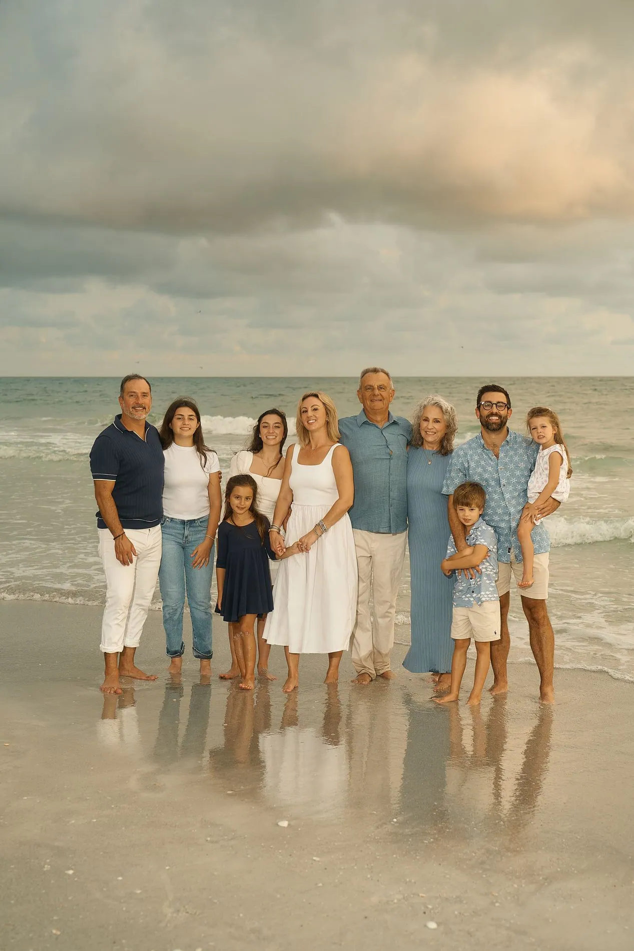 Ten people of three generations in white and blue standing barefoot in a row at the edge of the water under a cloudy sky, the grey-haired grandparents in the middle, a small girl on an arm at the end, their reflections in the wet sand