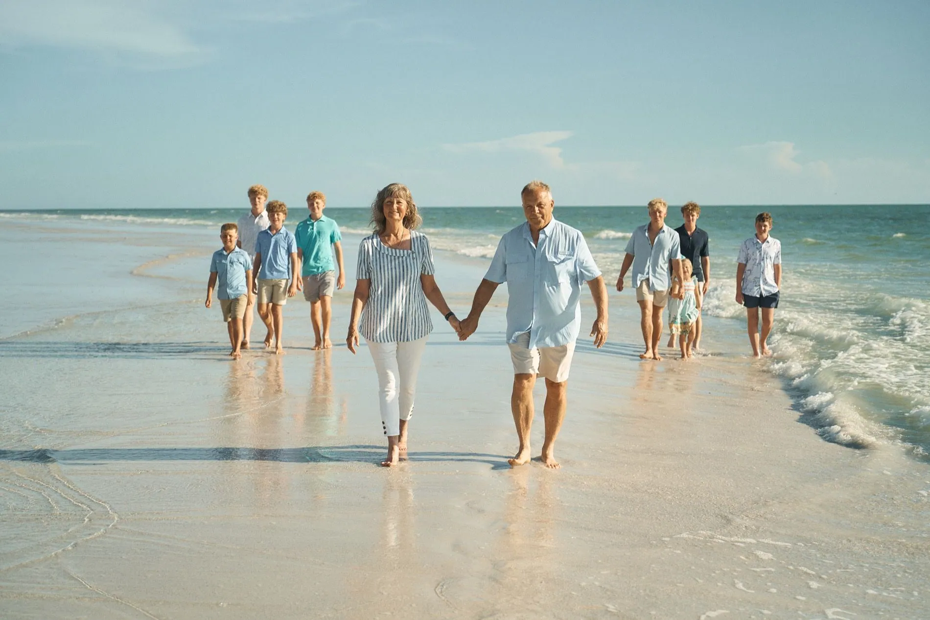 An older couple walking hand in hand along the edge of the water, the rest of their big family following behind them