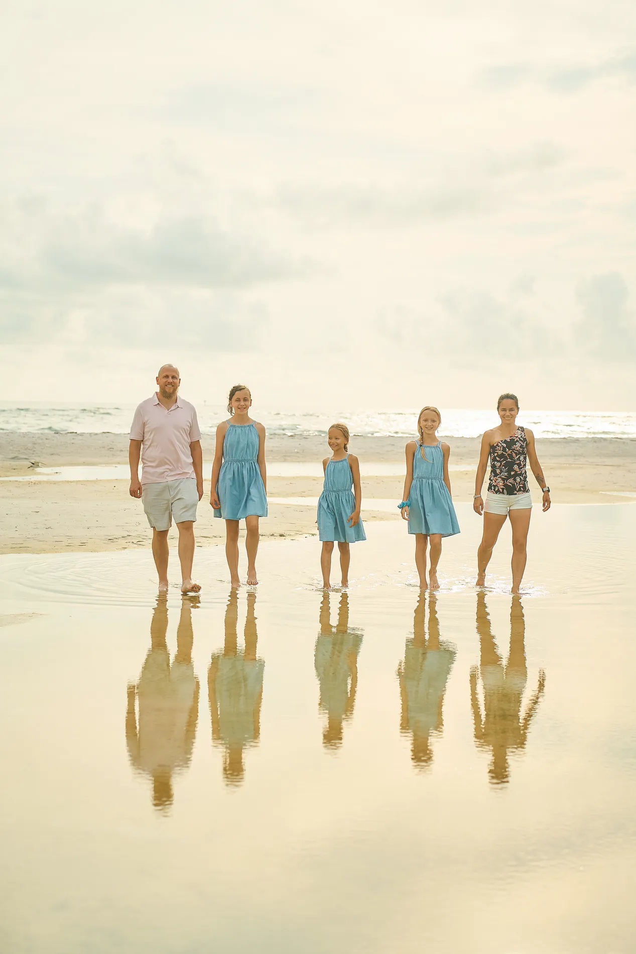 A family of five in blue and white standing in a row on wet sand on Lido Key, all reflected in a thin layer of water under a pale hazy sky