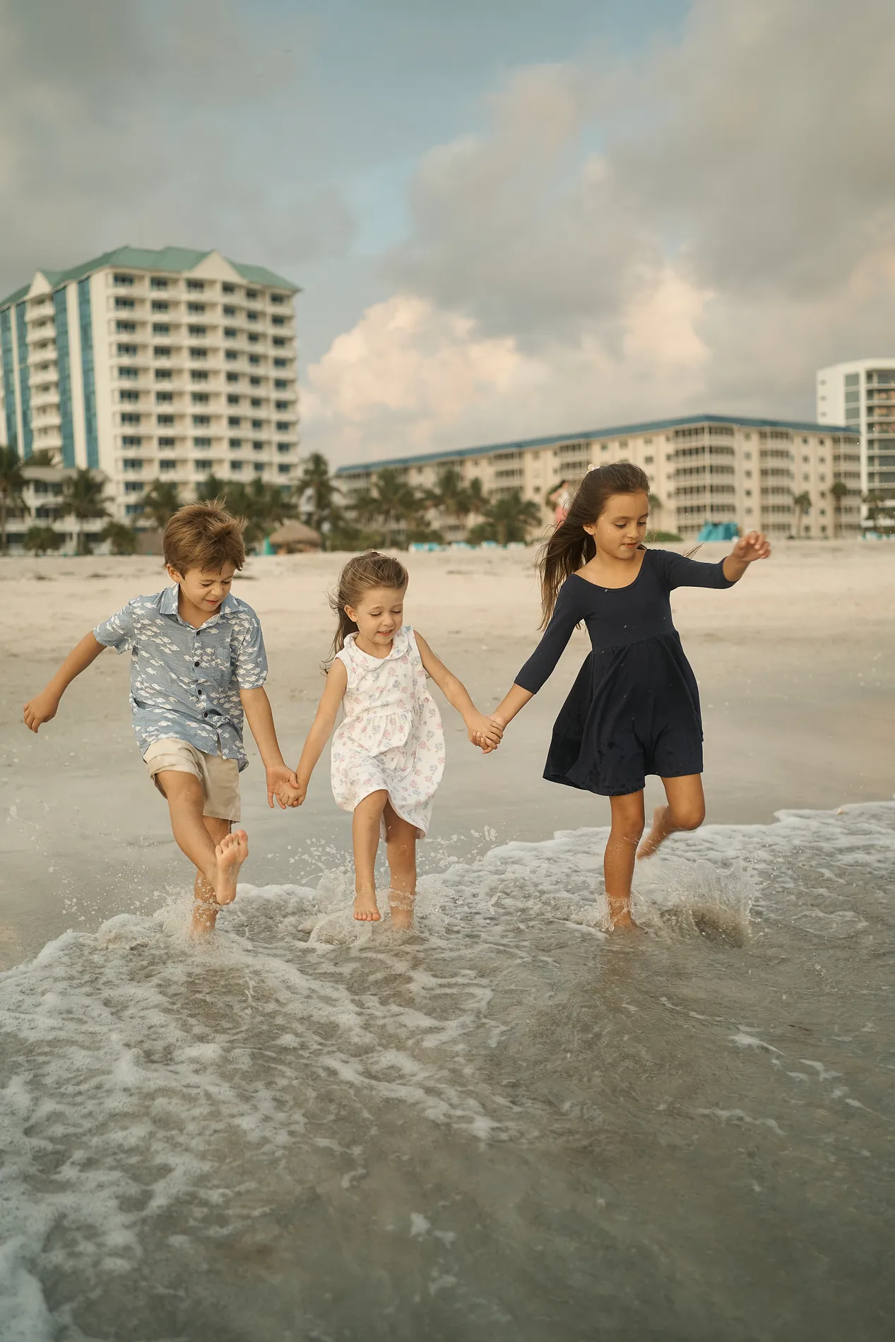 Three children holding hands and jumping over a small wave on Lido Key, a white hotel on the shore behind them