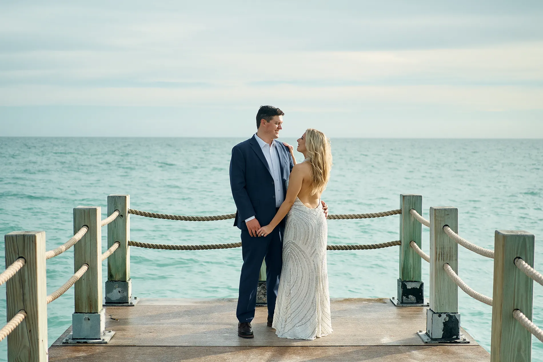 An engaged couple standing close on a wooden pier with rope railings, turquoise water all around them