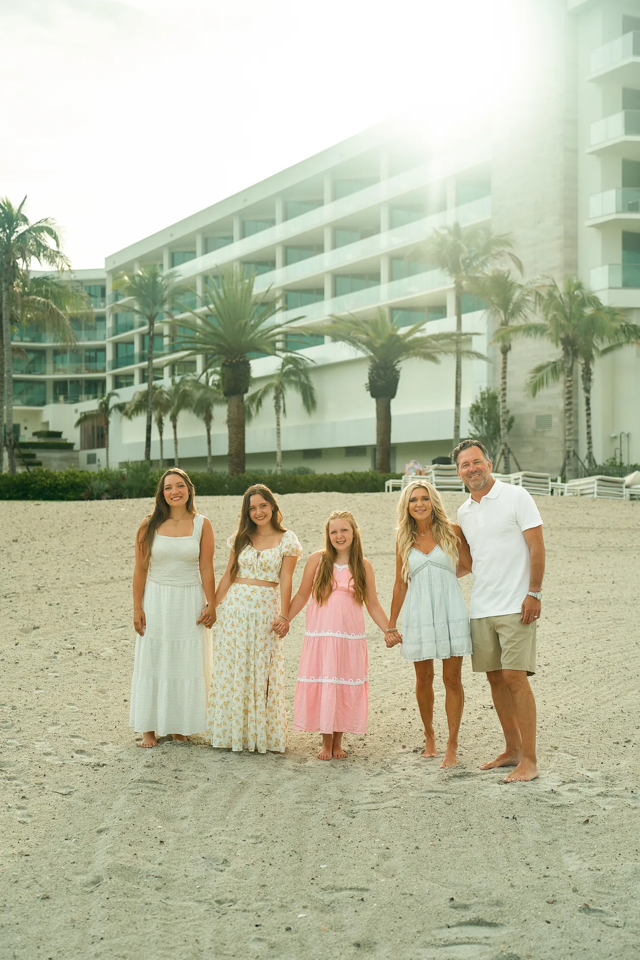 A father with his wife and three daughters in pastel dresses standing hand in hand on the sand of Longboat Key, palms and a beach hotel behind them
