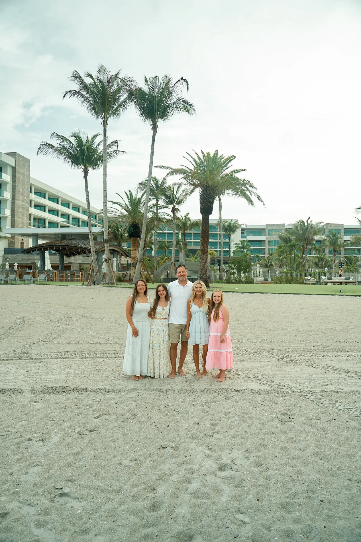 A family of five in white and pastel dresses standing together on the sand on Longboat Key under tall palms, a resort behind them and a flat white overcast sky