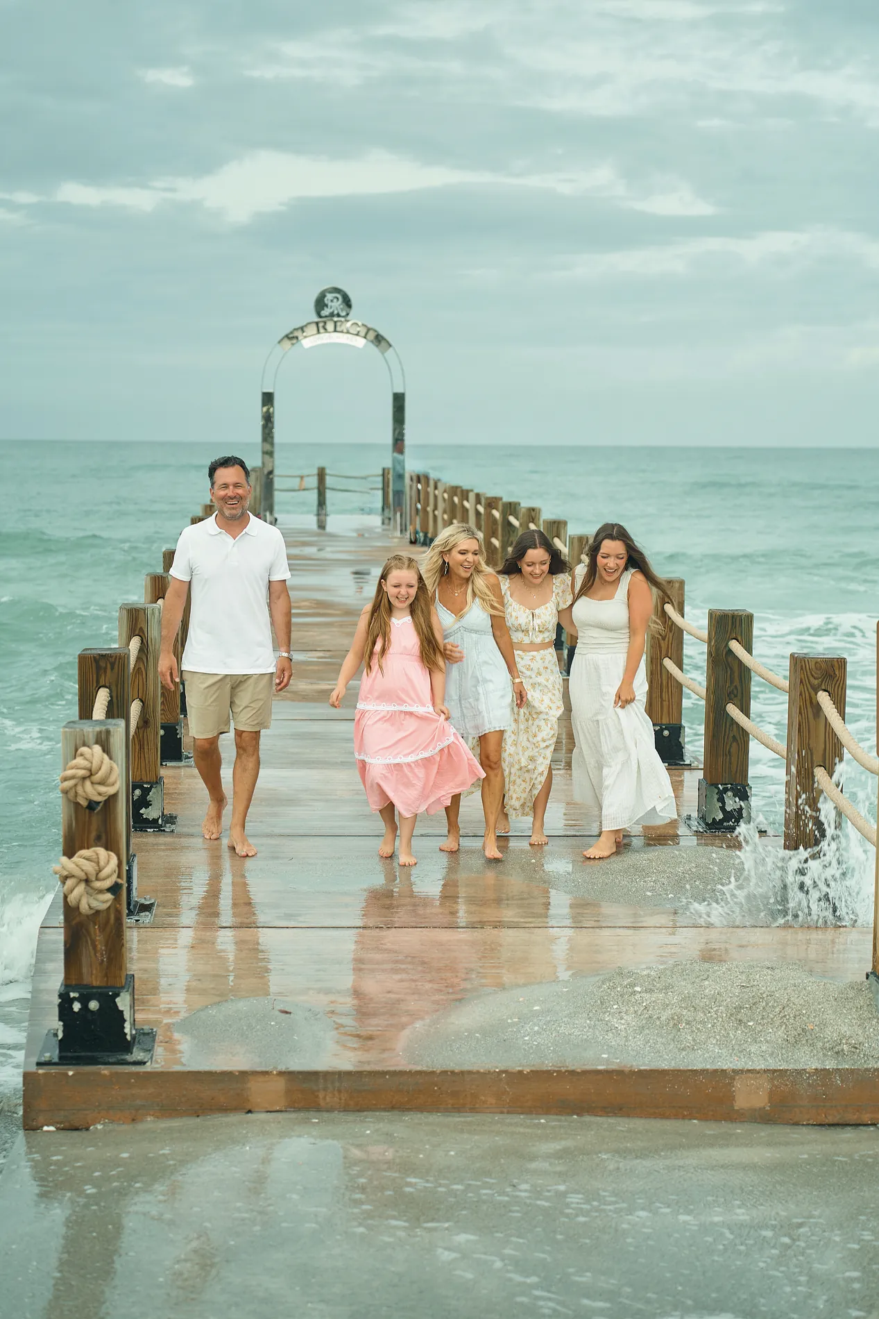 A father in white, a girl in a pink dress and three women in light dresses walking toward the camera on a wooden pier on Longboat Key, a wave splashing against the posts and clouds over turquoise water
