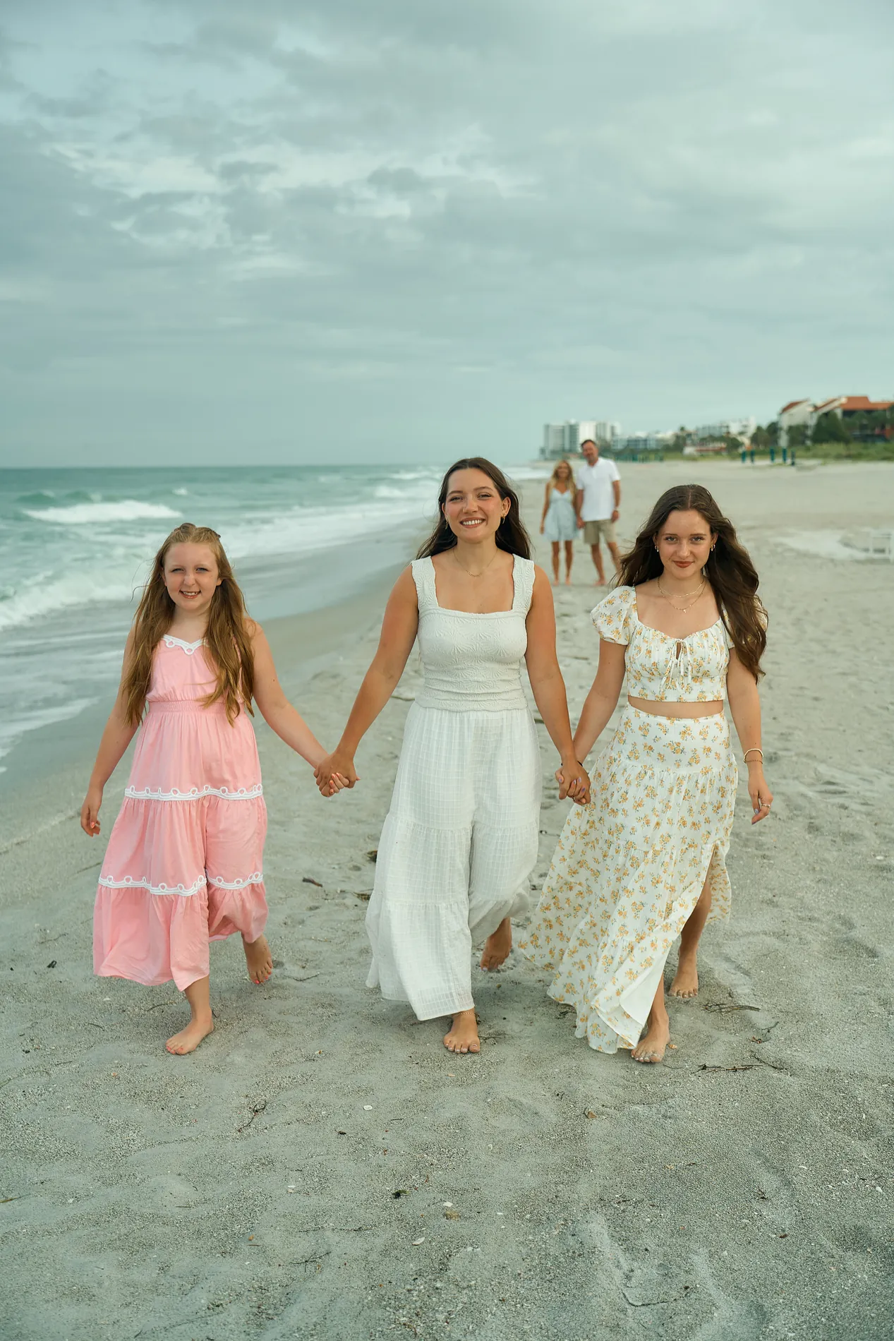 Three young women in long dresses walking hand in hand toward the camera along the water on Longboat Key