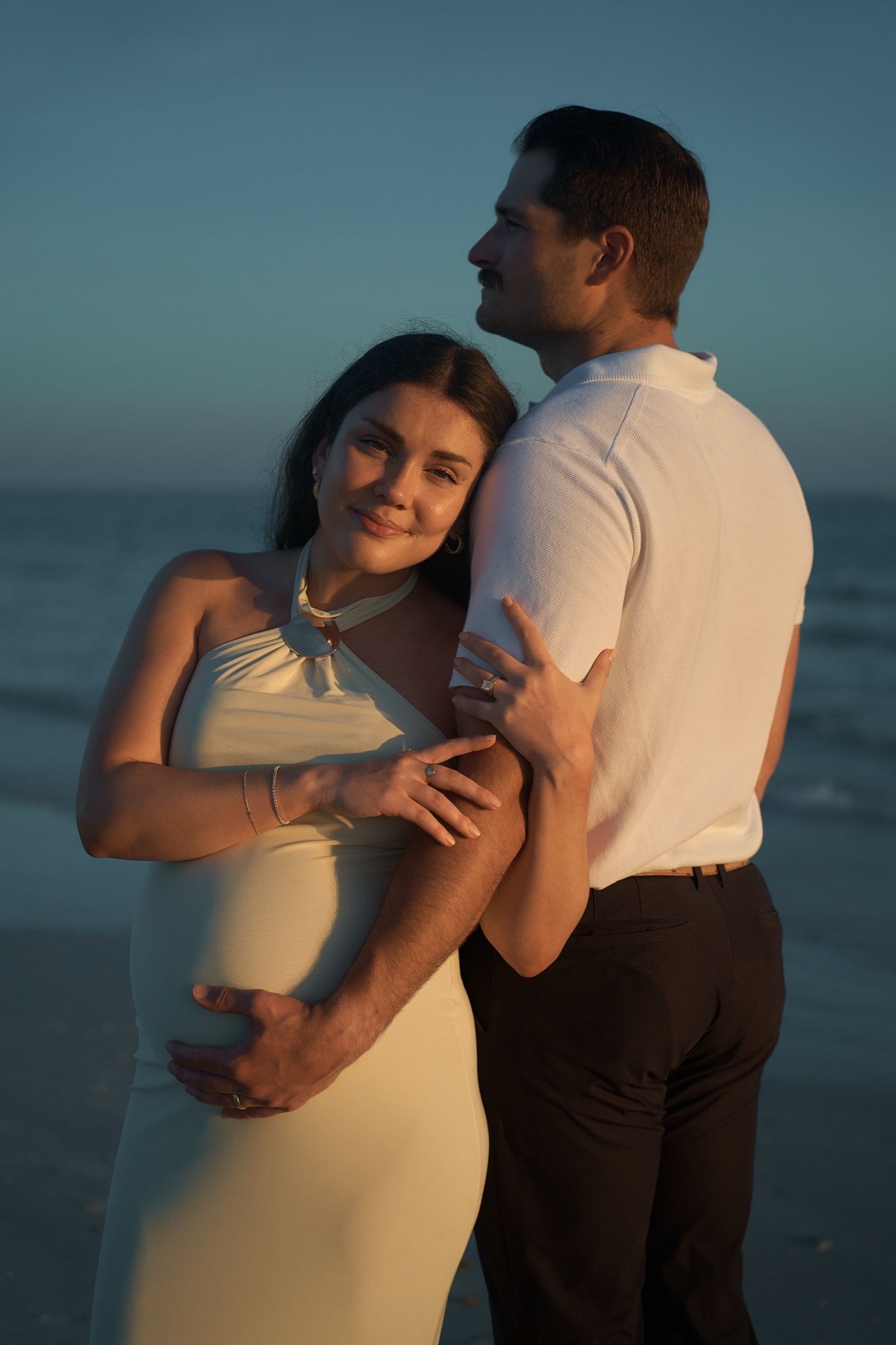 A couple on the beach at dusk, her hands on her belly, the Gulf behind them