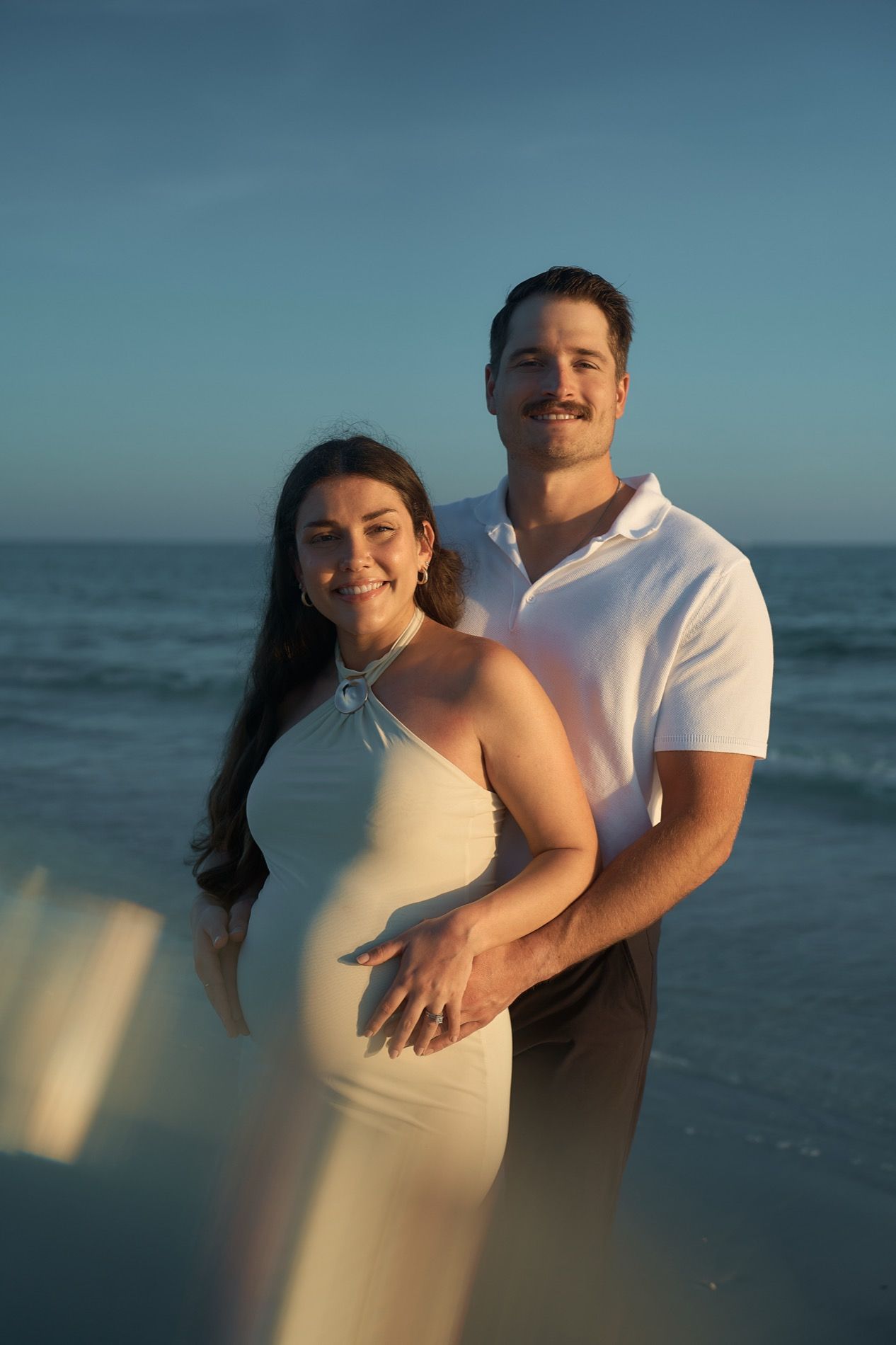A pregnant woman and her partner standing at the water on a Sarasota beach in the last sunlight