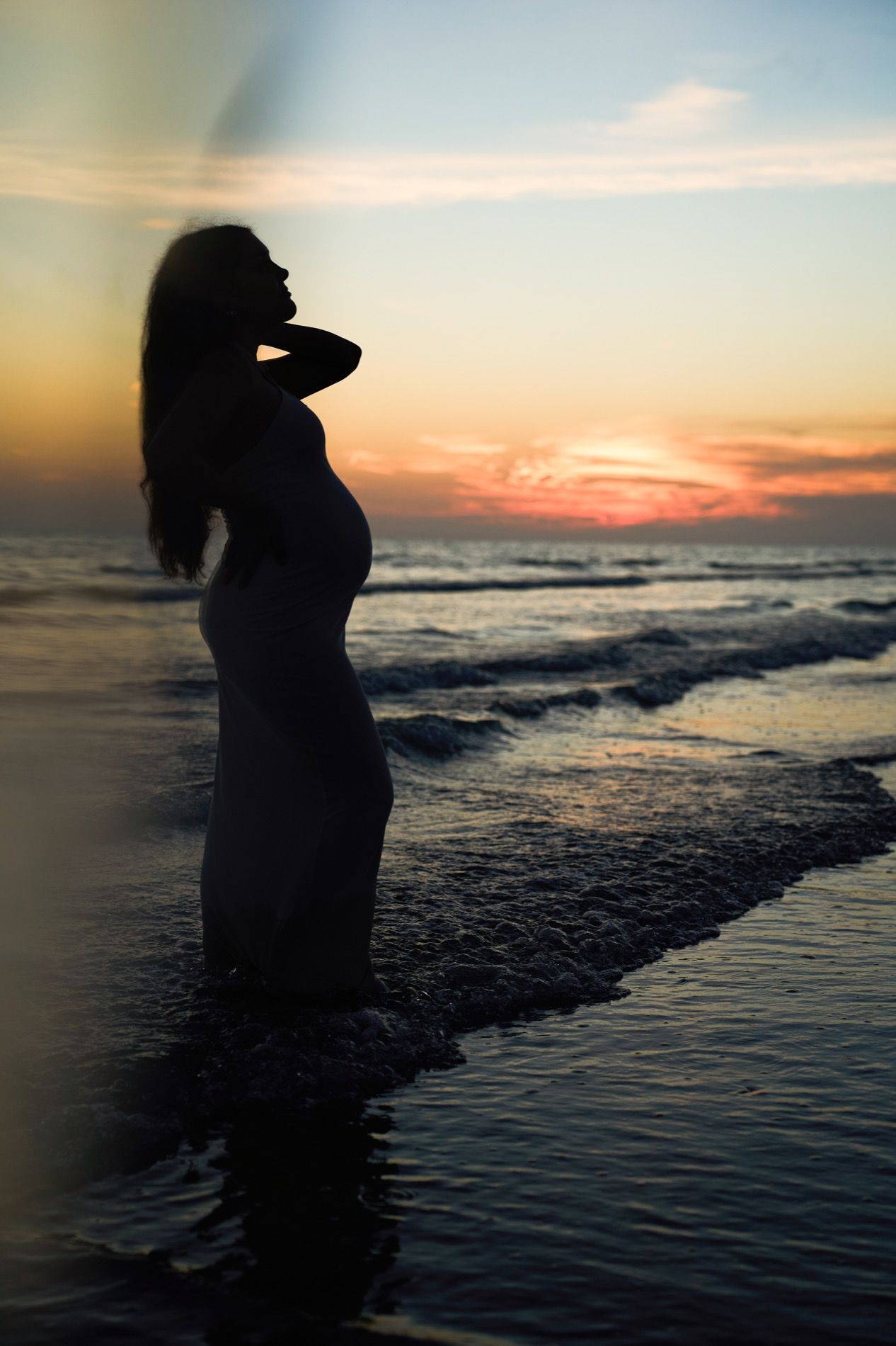 A silhouette of a pregnant woman at the shoreline after sunset, the last light of the sky behind her