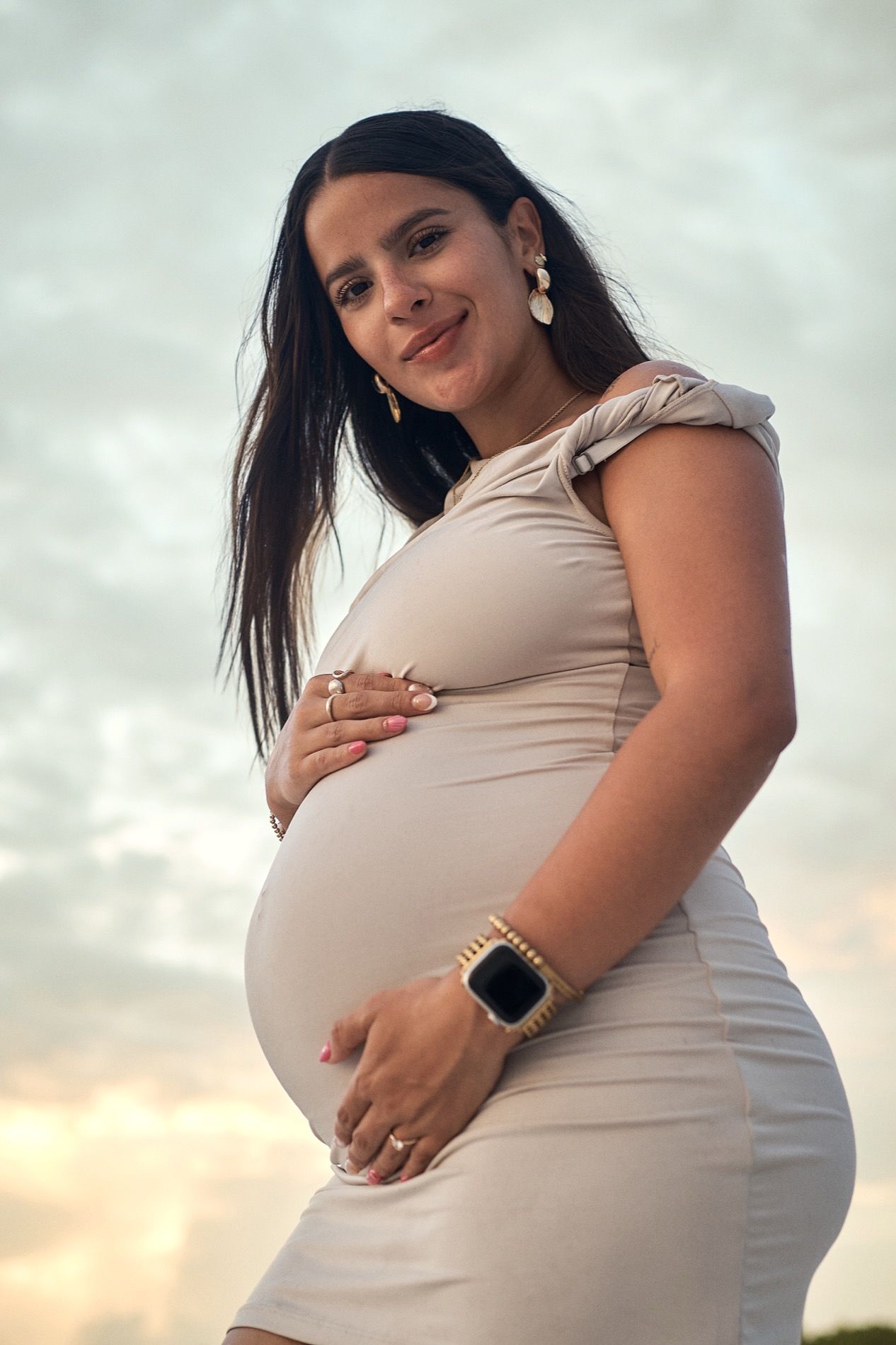 A pregnant woman in a fitted dress holding her belly on a Sarasota beach under a soft evening sky