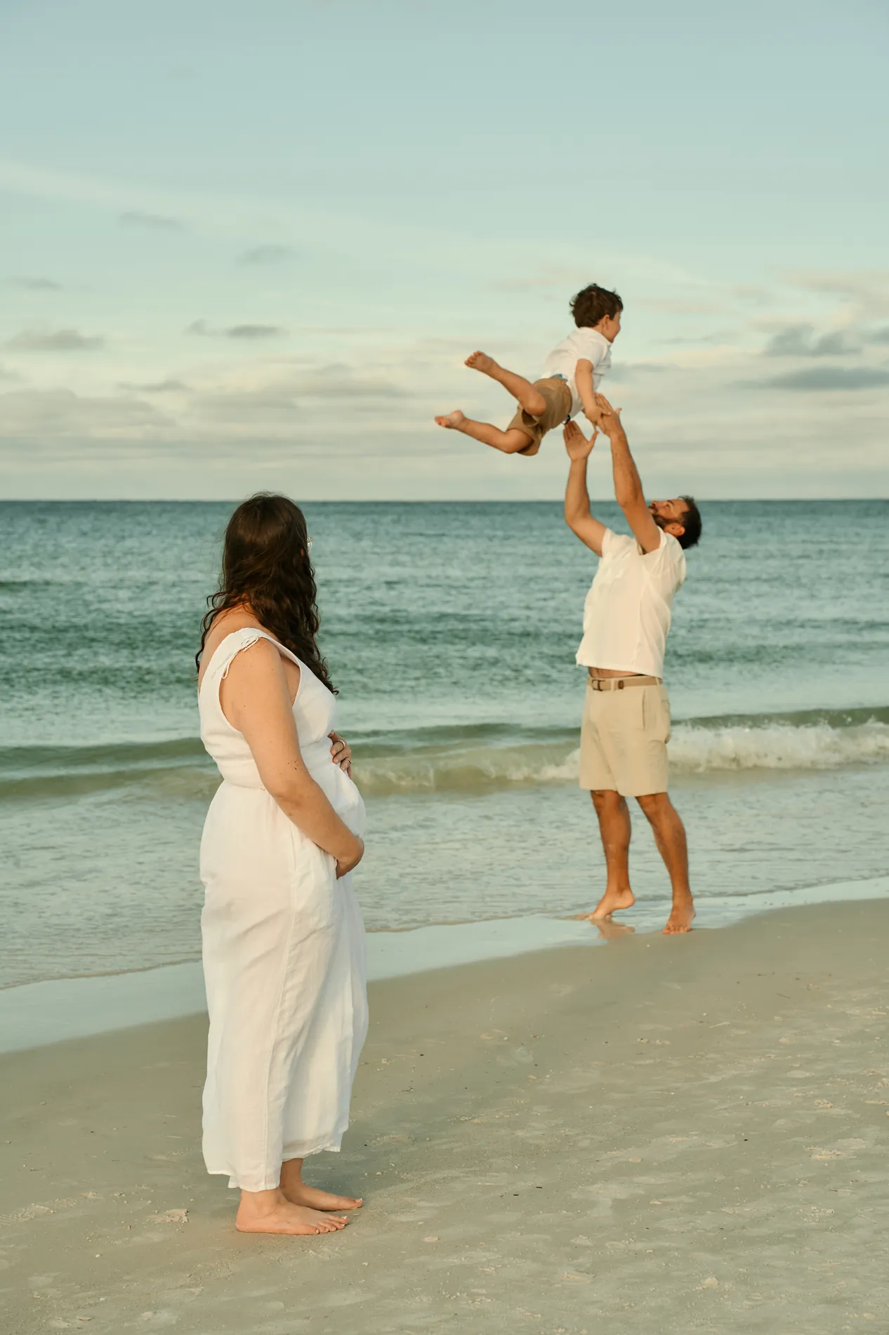 A pregnant mother in a long white dress standing at the water, watching the father toss their young son into the air above the Gulf
