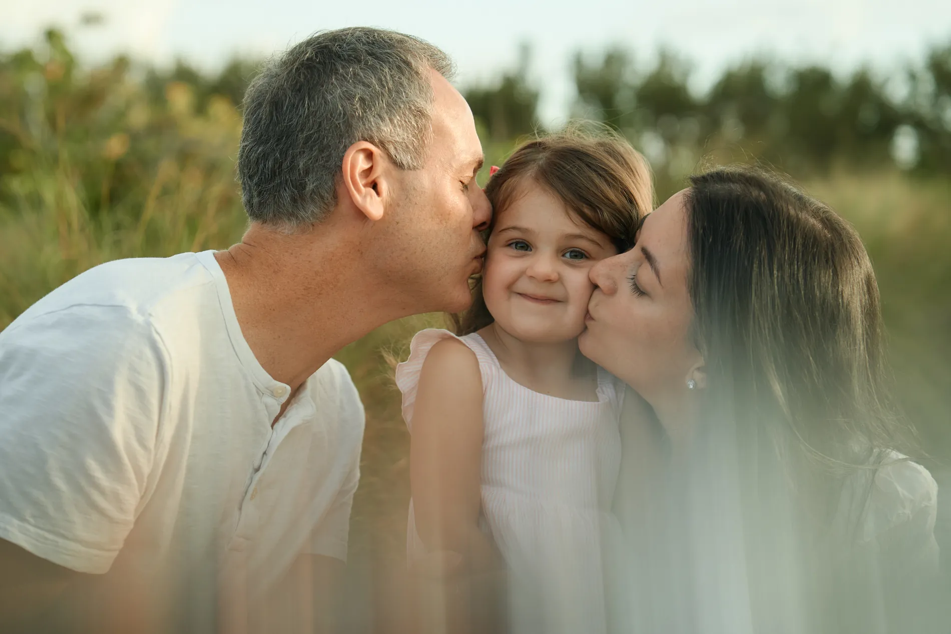 A father and mother kissing their little daughter on both cheeks among the dune grass on Siesta Key, the girl smiling at the camera