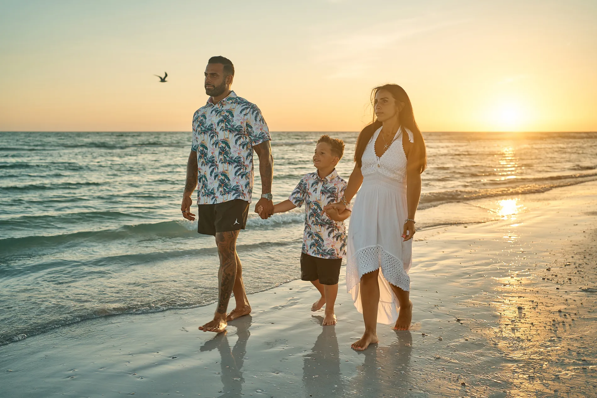 A father, a young son and a mother in white walking hand in hand on the wet sand as the sun sets into the Gulf