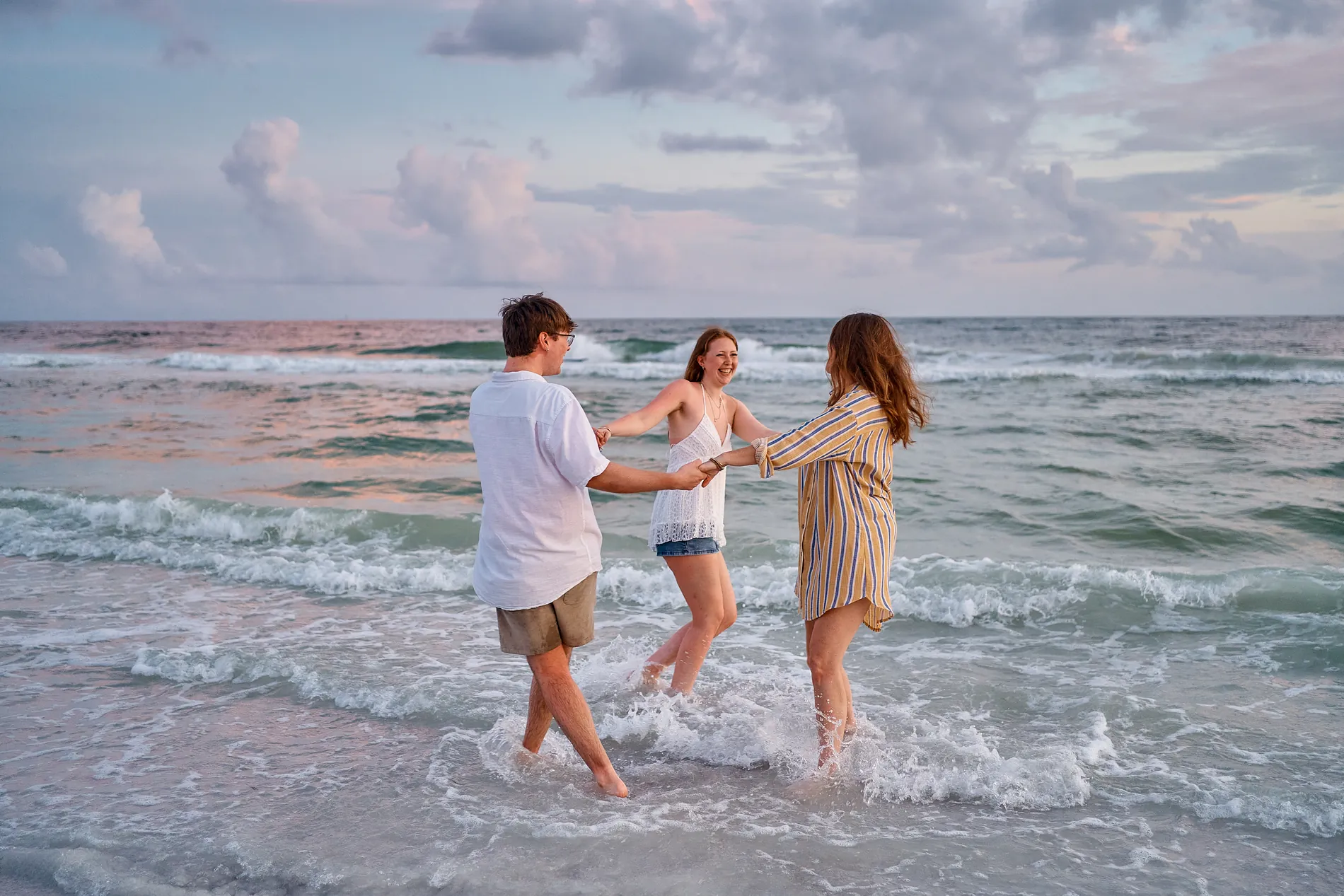 Three people laughing and splashing in the shallow water under a soft lilac evening sky
