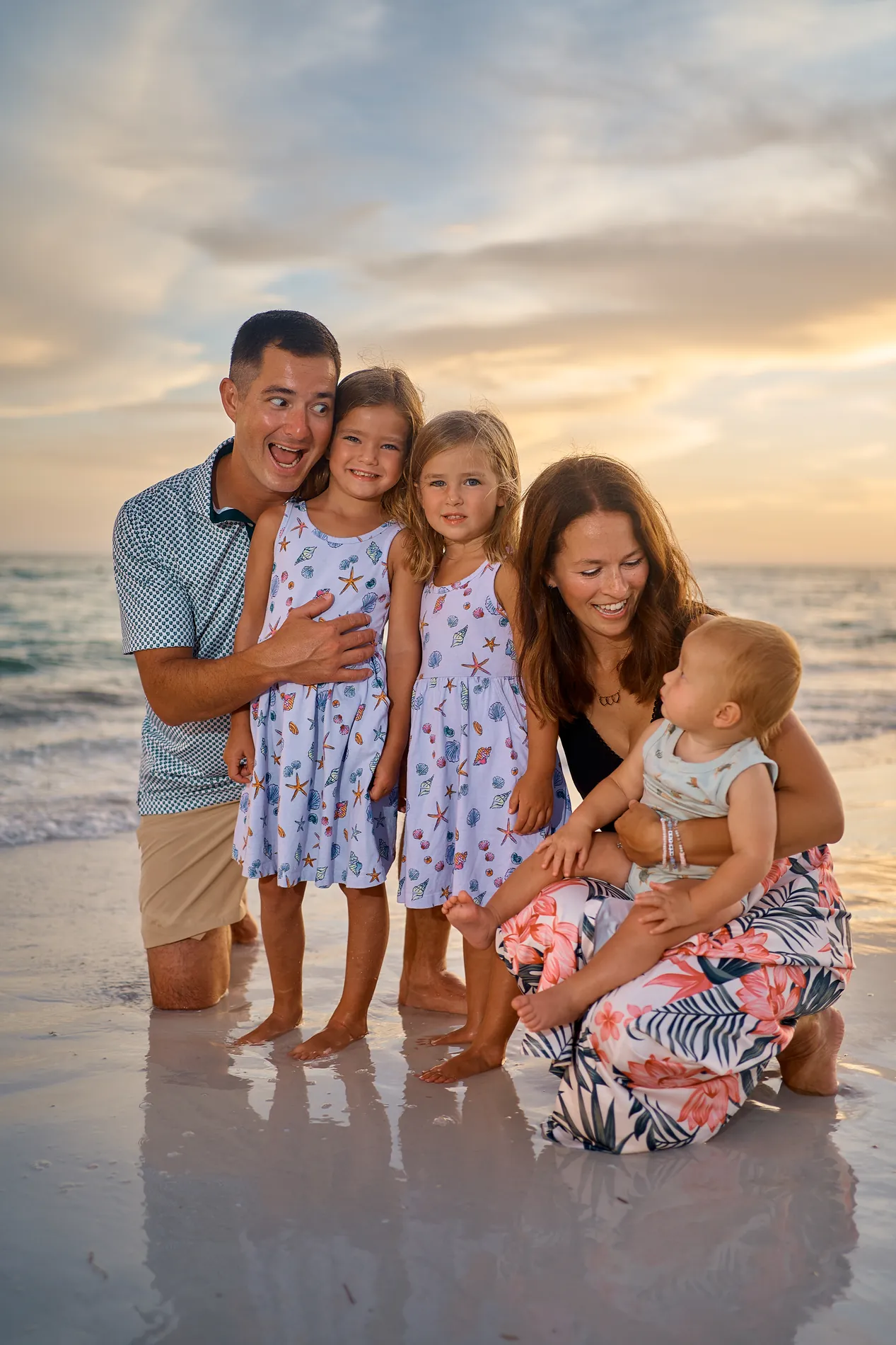 A father kneeling at the water line on Siesta Key with two little daughters in matching floral dresses, the mother kneeling beside them with the baby, under a sunset sky