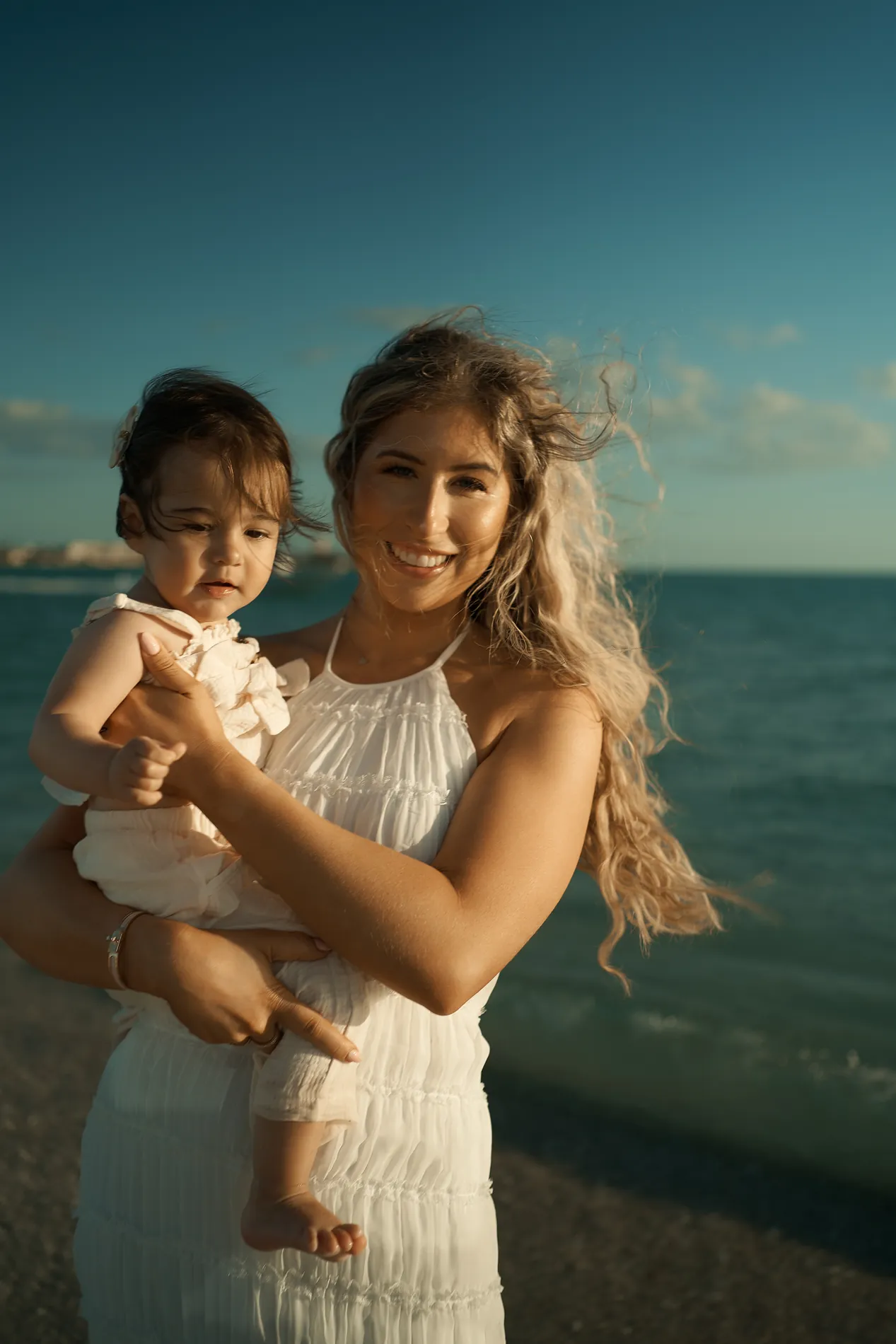 A smiling mother in a white sundress holding her baby at the edge of the sea, her long fair hair blown sideways by the wind, dark turquoise water behind them