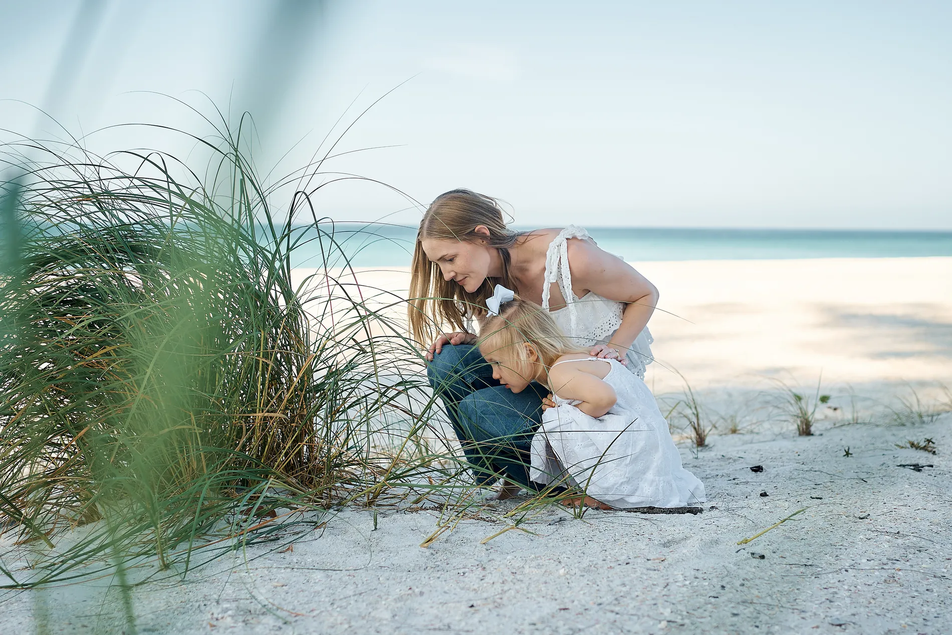 A mother crouching with her little daughter beside a clump of sea oats, turquoise water and an almost empty white beach behind them