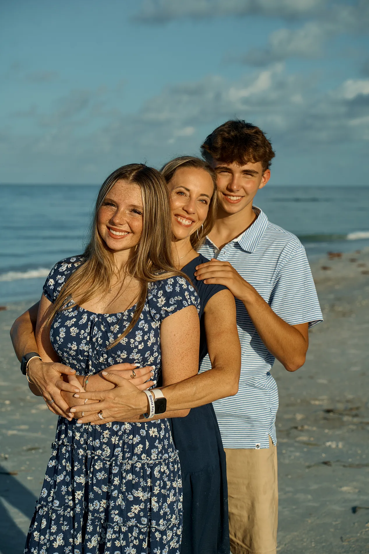 A mother hugged by her teenage daughter in a floral dress and her teenage son at the edge of the water, a blue evening sky behind them