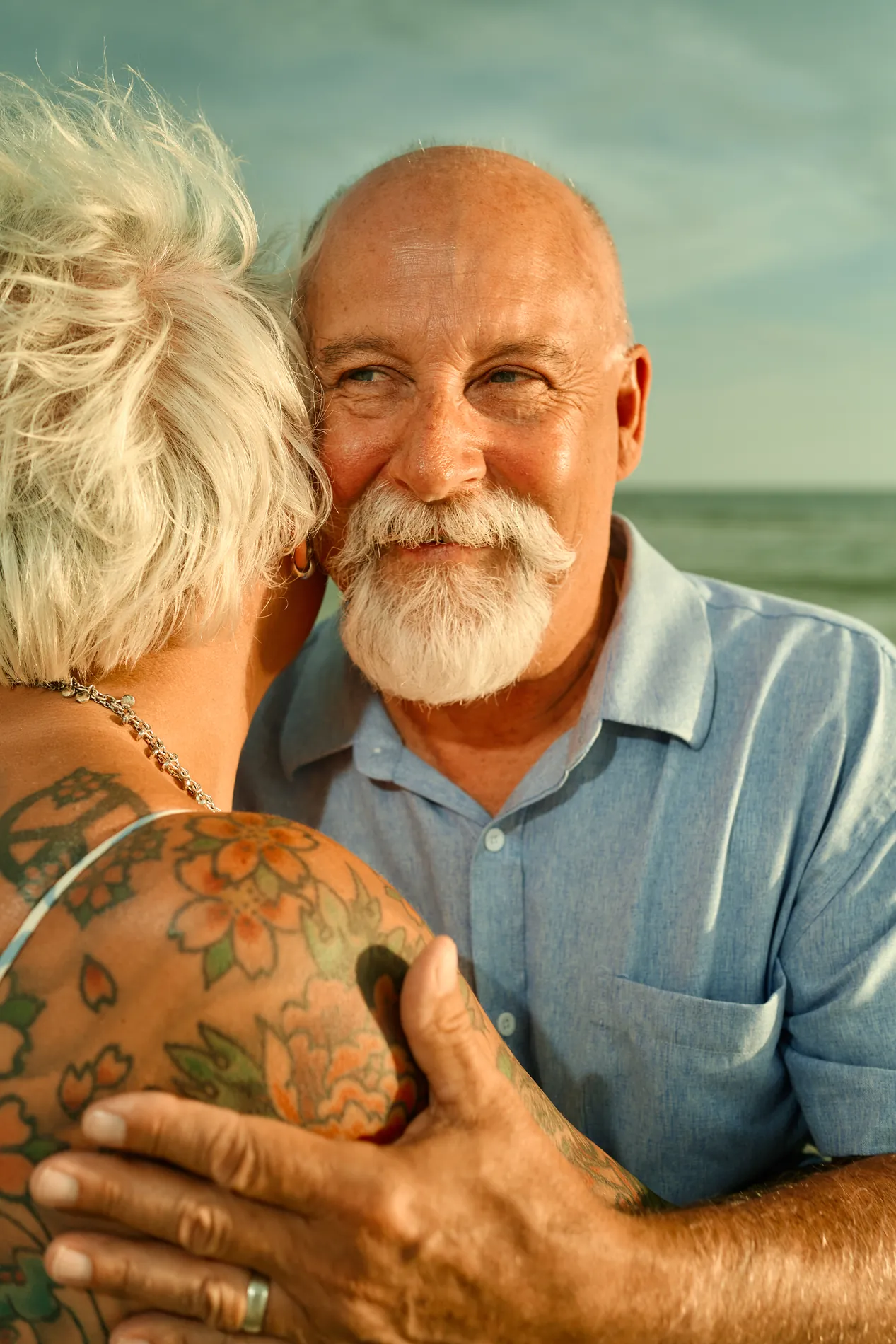 An older couple close up at the water: a man with a white beard smiling as he holds his wife, his hand with a wedding ring on her shoulder