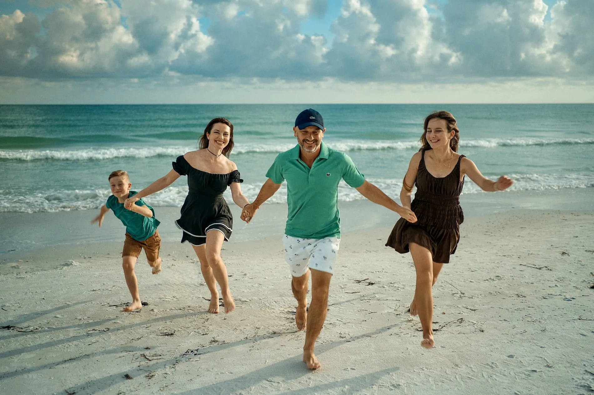 A family of four running hand in hand along the waterline toward the camera under a sky of big white clouds