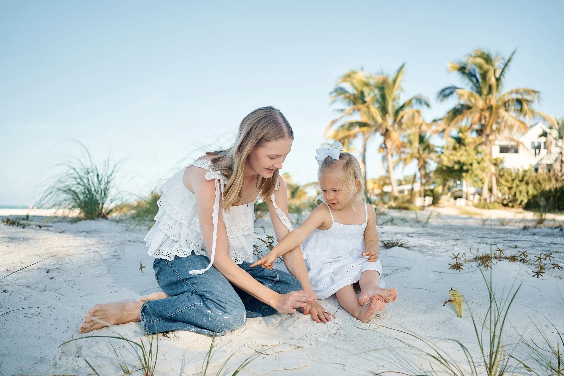 A mother in white kneeling on the sand beside her little daughter in a white dress, sea grass and palm trees behind them