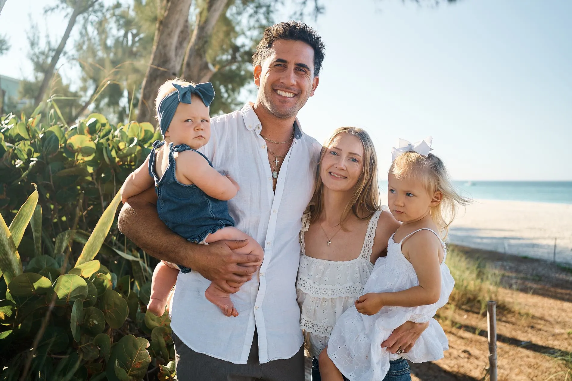 A father holding a toddler girl in a blue dress, the mother and an older daughter in white beside them, all smiling on a beach with palms behind