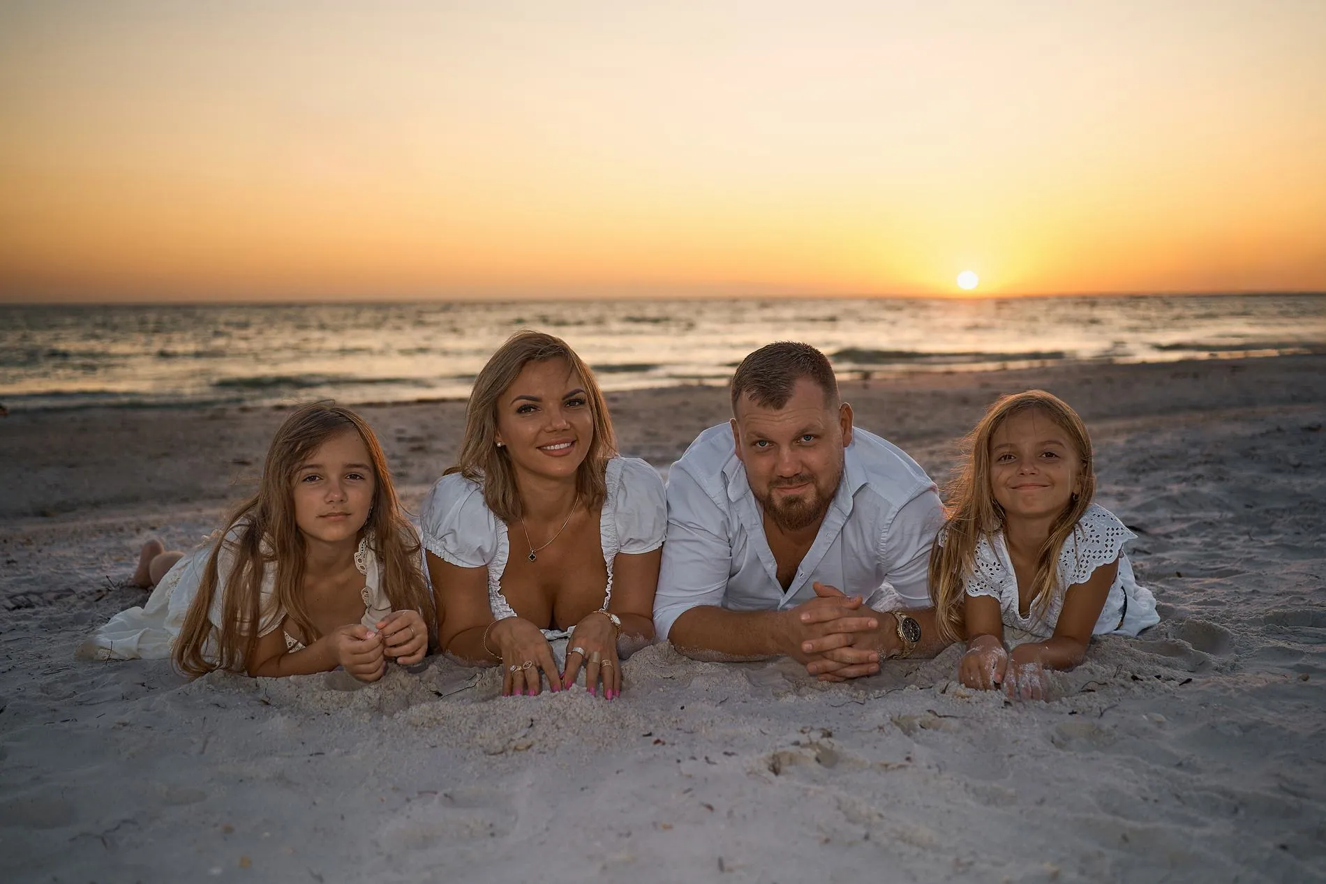 A father, a mother and their two daughters lying on the sand in a row, chins on their hands, the sun setting over the sea behind them