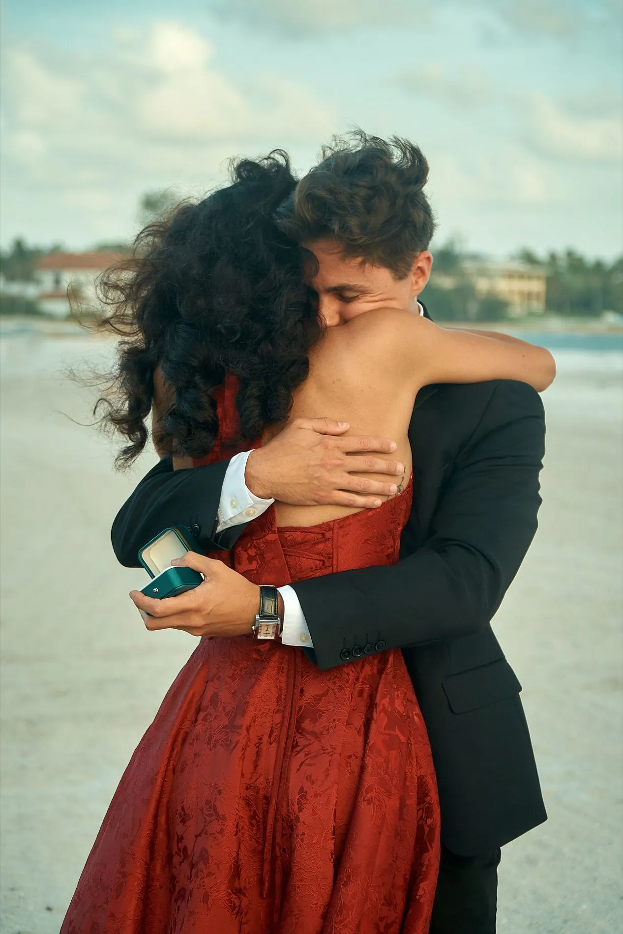 A woman in a red dress holding her new fiance tight in a hug, an open ring box in his hand behind her back