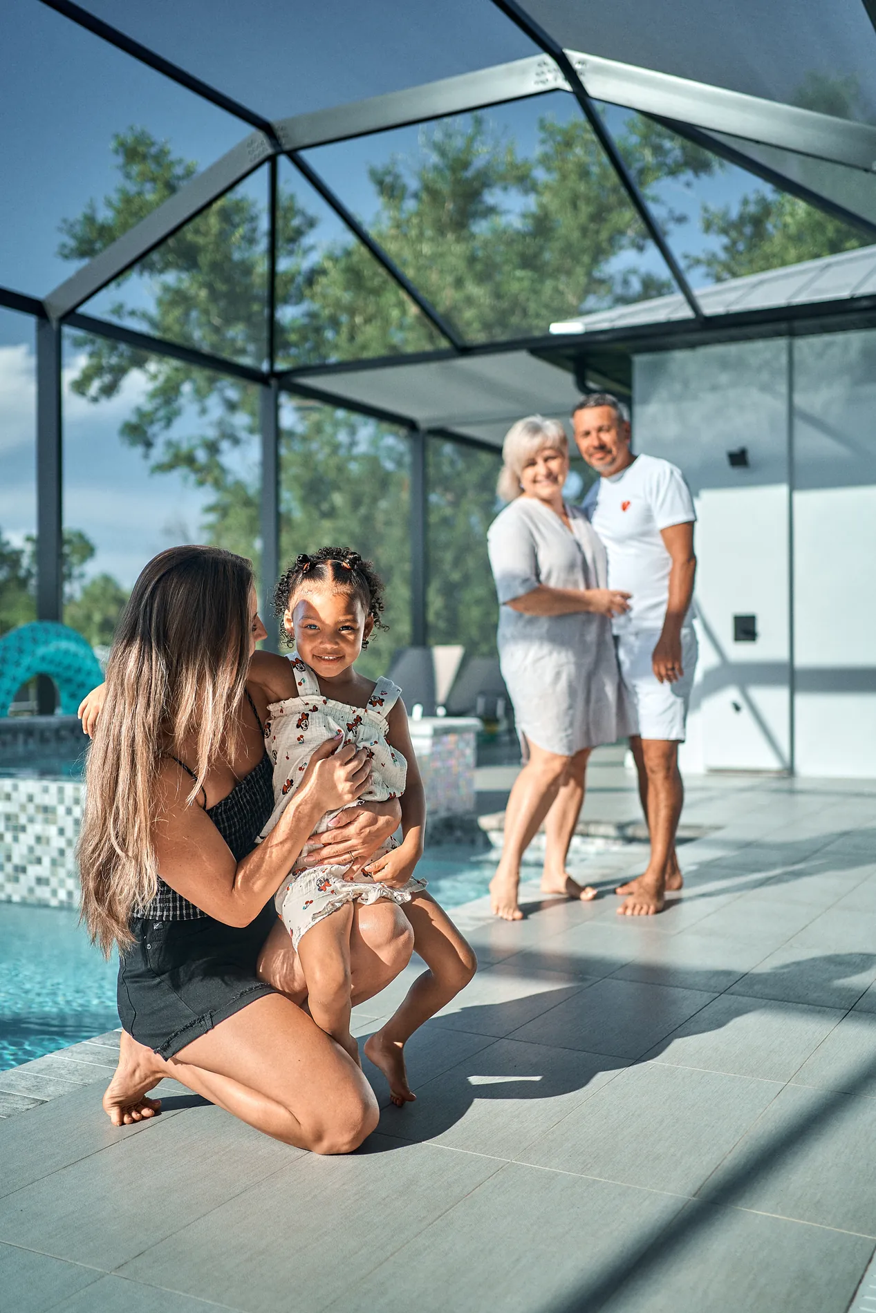 A woman crouching to hug a laughing little girl beside the pool on a screened lanai, an older couple standing behind them with their arms around each other, watching