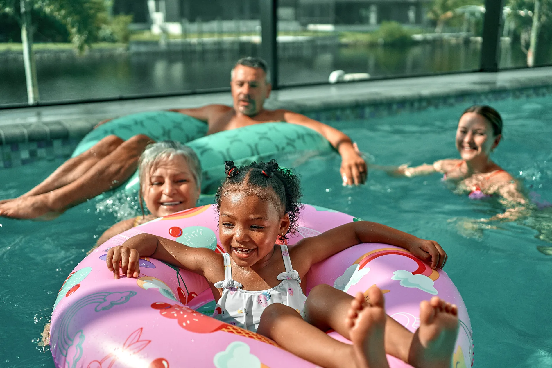 A little girl laughing in a pink ring float in a screened pool, an older couple and a young woman floating and smiling behind her