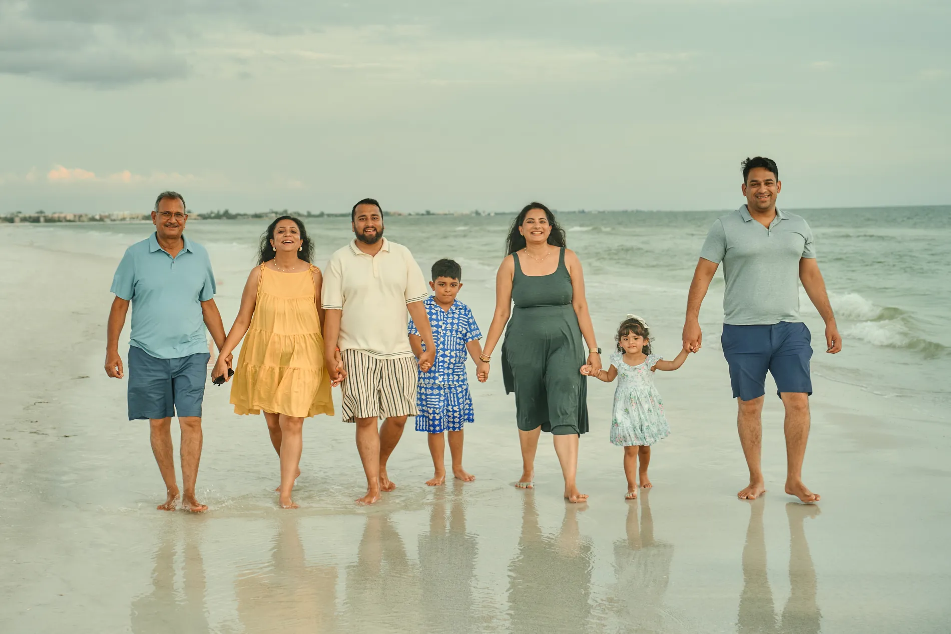 Three generations of one family, seven people, walking hand in hand through the shallow water toward the camera, the grandfather on the left and a little girl in a flowered dress near the right