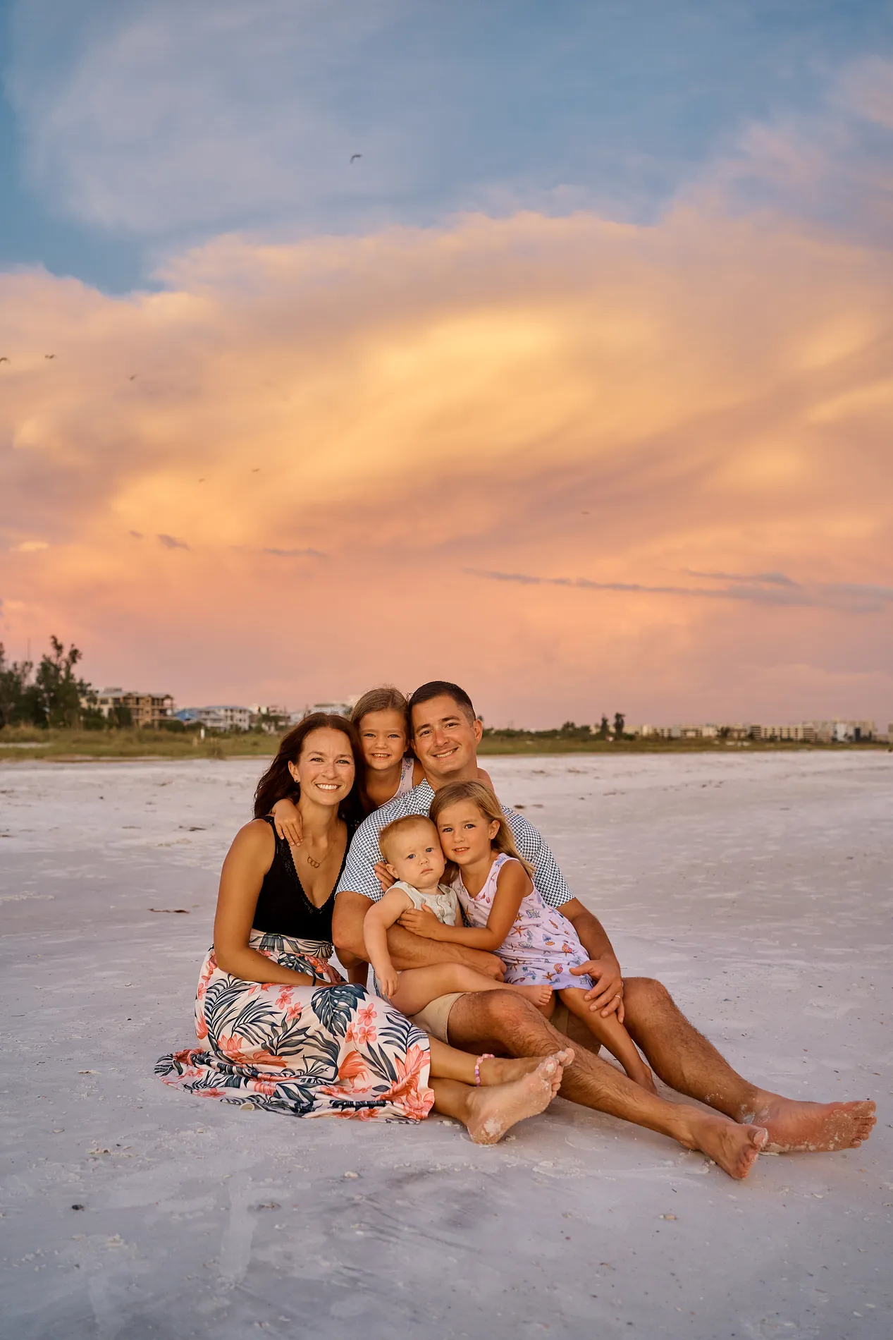 A family of five sitting close together on the white sand of Siesta Key, the parents holding a baby and two little girls in matching floral dresses, pink and orange clouds above them after sunset
