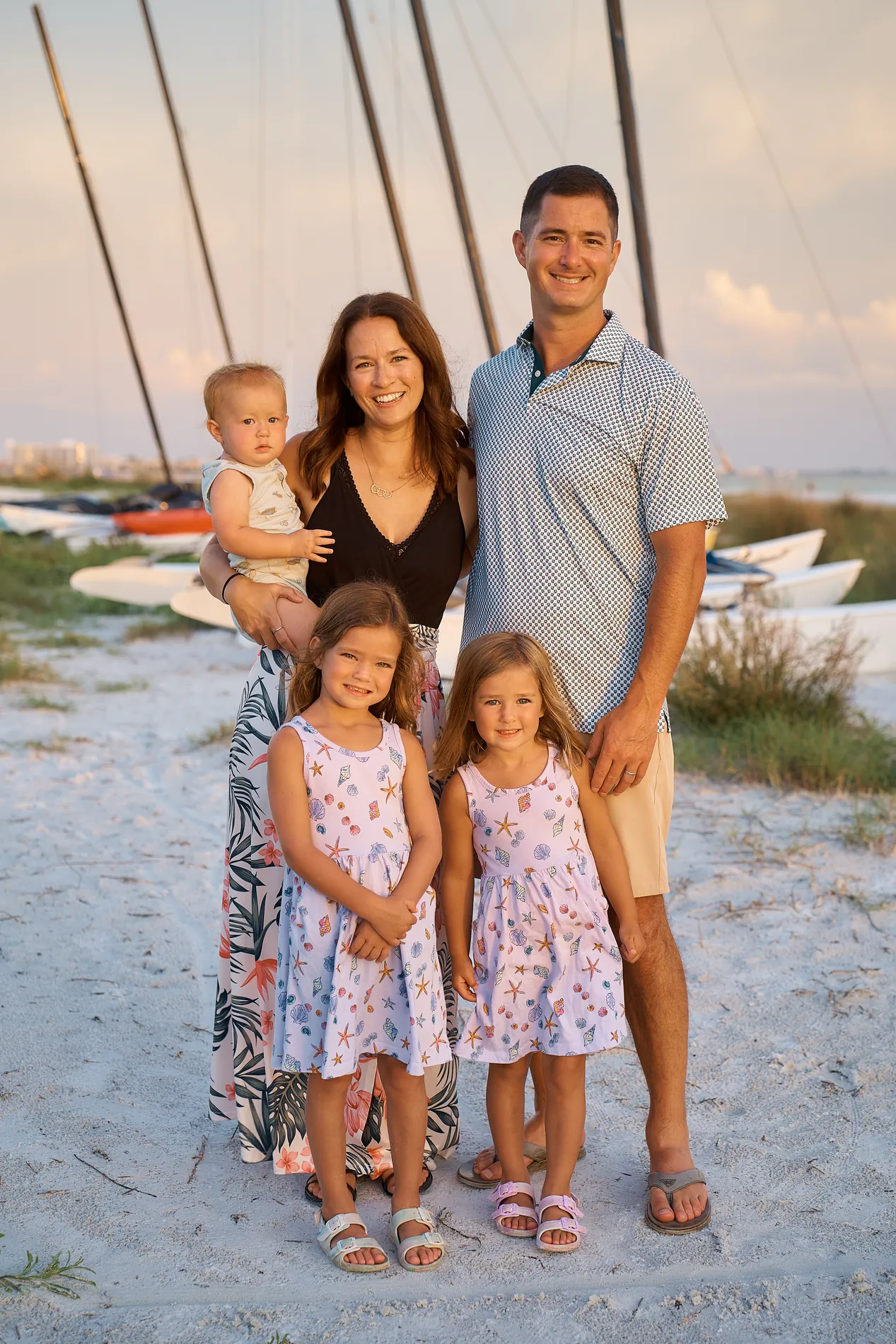 A mother holding a baby, a father and two little girls in matching floral dresses standing on Siesta Key beach, catamaran masts behind them in soft evening light