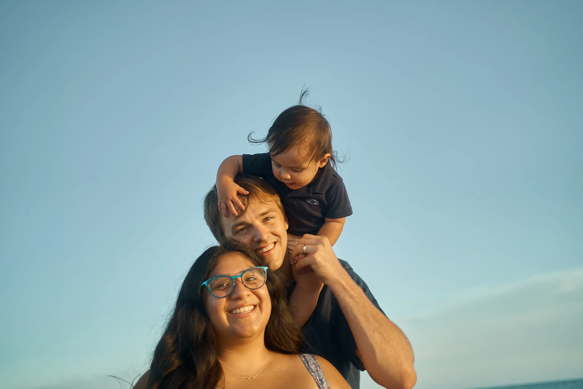 A smiling mother in glasses beside a father carrying their toddler son on his shoulders on Siesta Key, a clear blue sky behind them