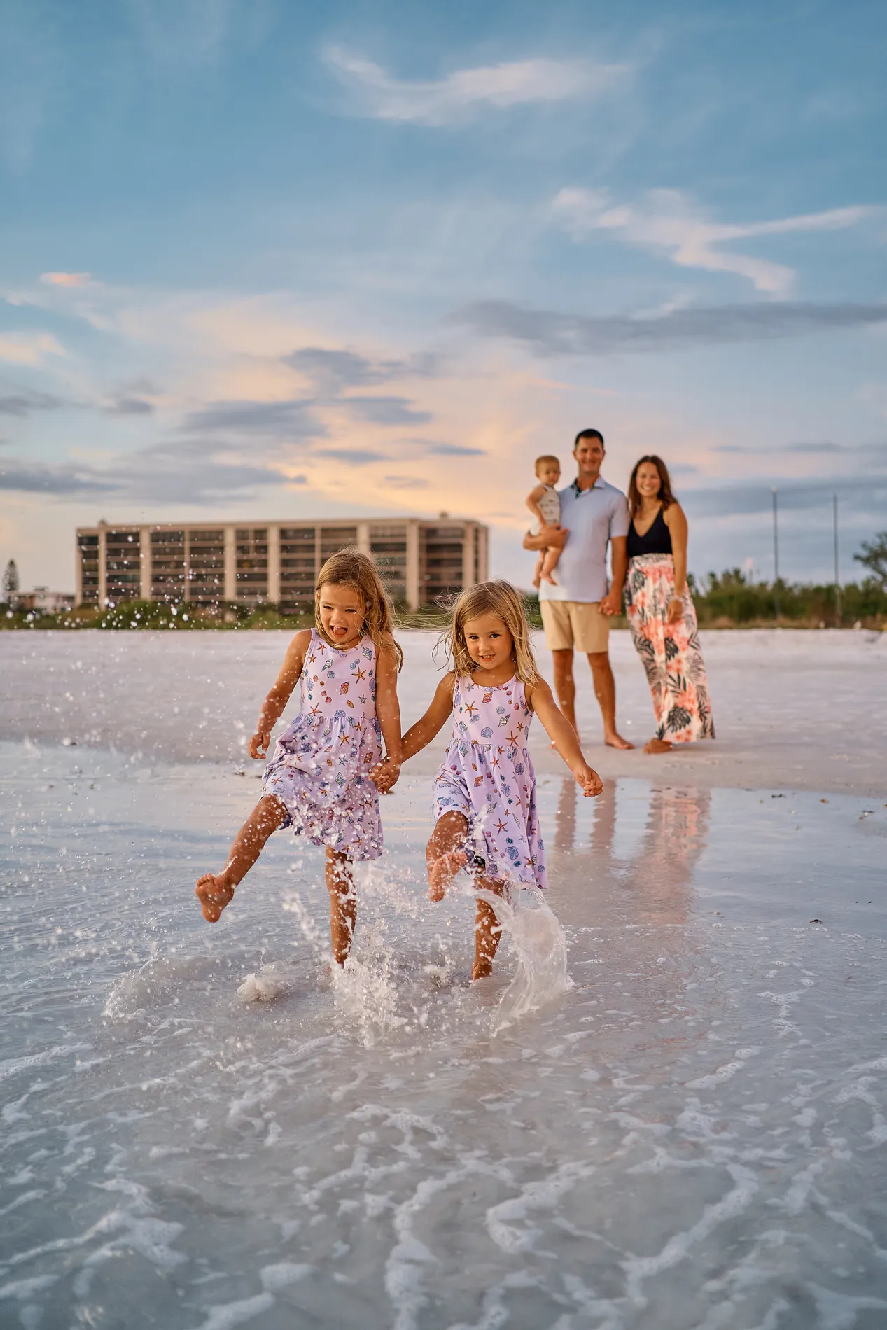 Two little girls in matching dresses running and splashing through the shallow water on Siesta Key, their parents holding a baby behind them