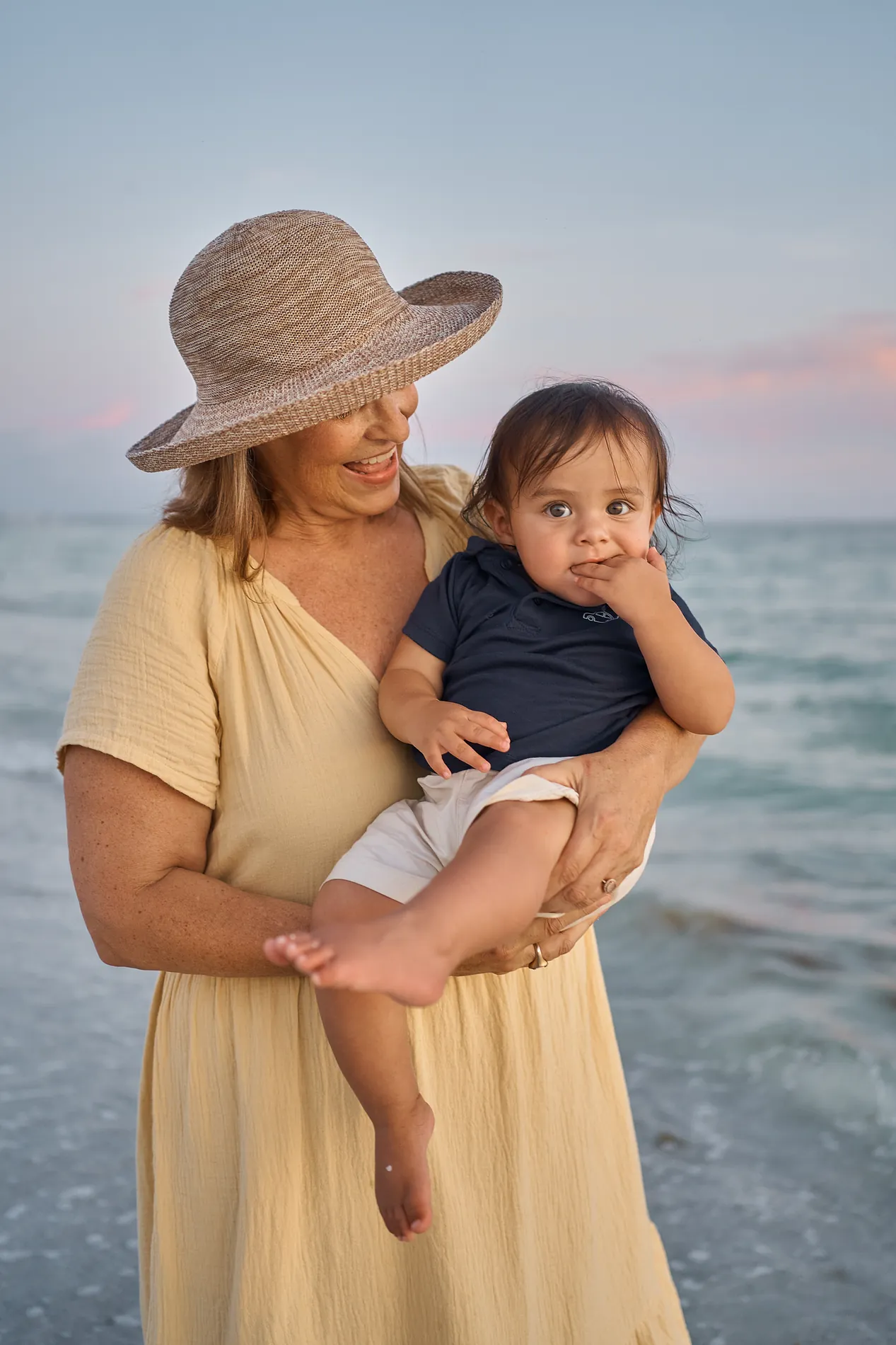 A laughing grandmother in a straw hat holding her grandson at the water on Siesta Key, the sky pink and lilac just after sunset