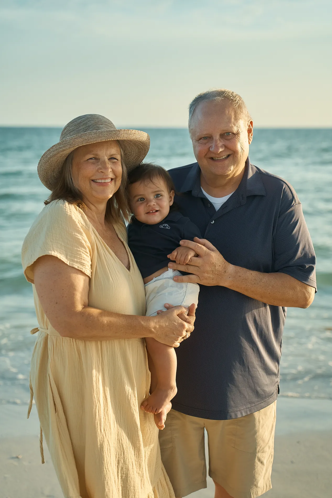 A grandmother in a straw hat and a grandfather holding their baby grandson between them in front of turquoise water on Siesta Key