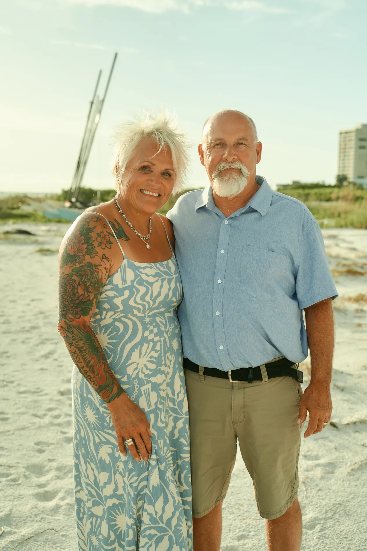 A smiling grandmother in a blue and white patterned maxi dress and a grandfather in a light blue shirt and khaki shorts standing together on Siesta Key, catamaran masts behind them