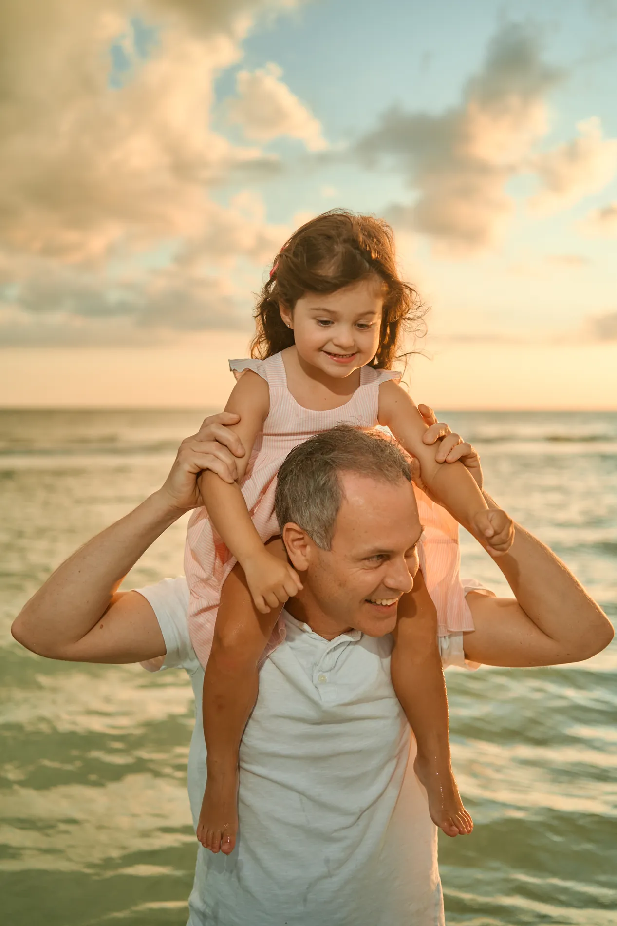 A grey-haired father standing in the sea on Siesta Key with a laughing little girl in pink on his shoulders, a warm evening sky behind them