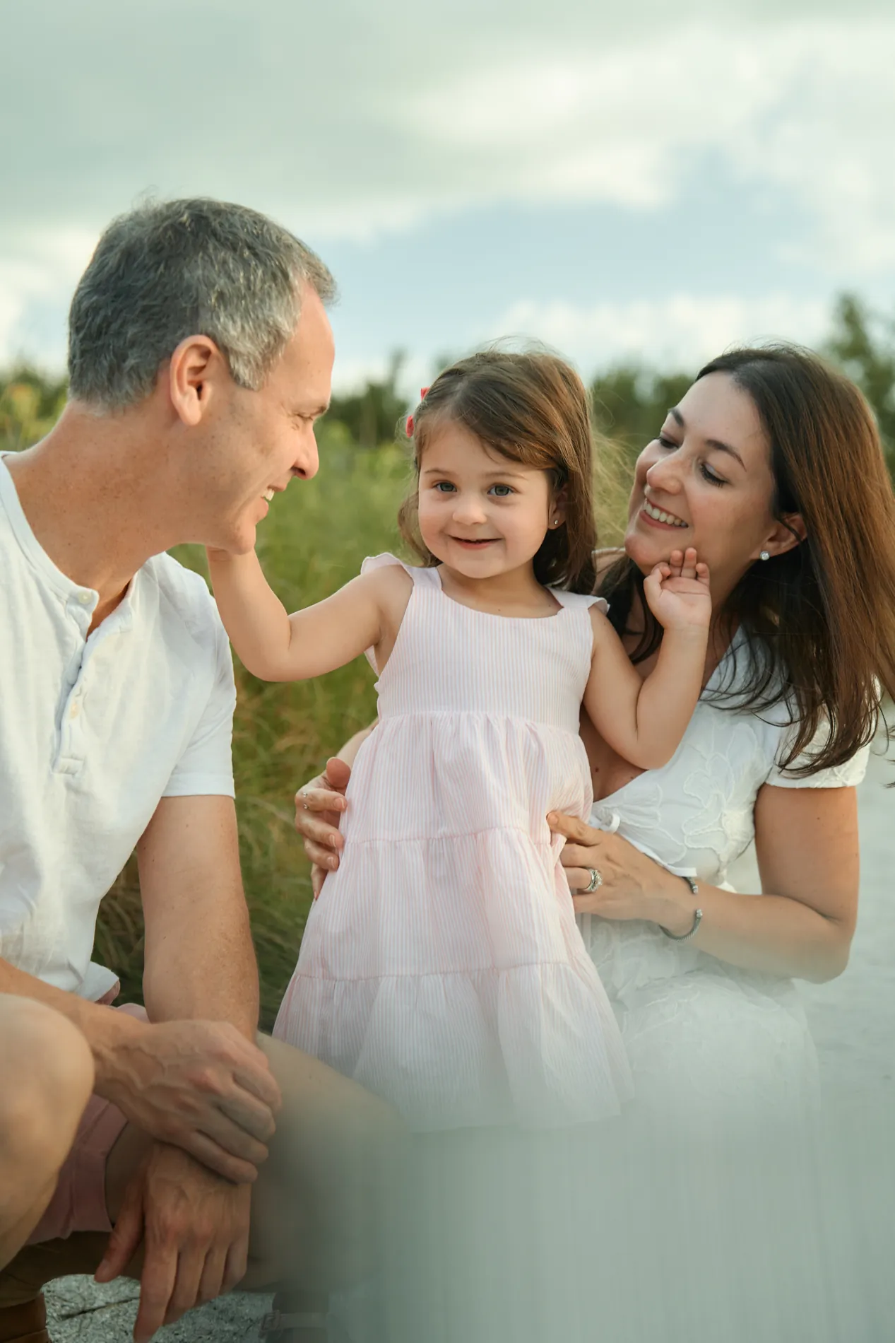 A grey-haired man and a woman crouching on the sand beside a smiling little girl in a pink dress on Siesta Key, dune grass behind them