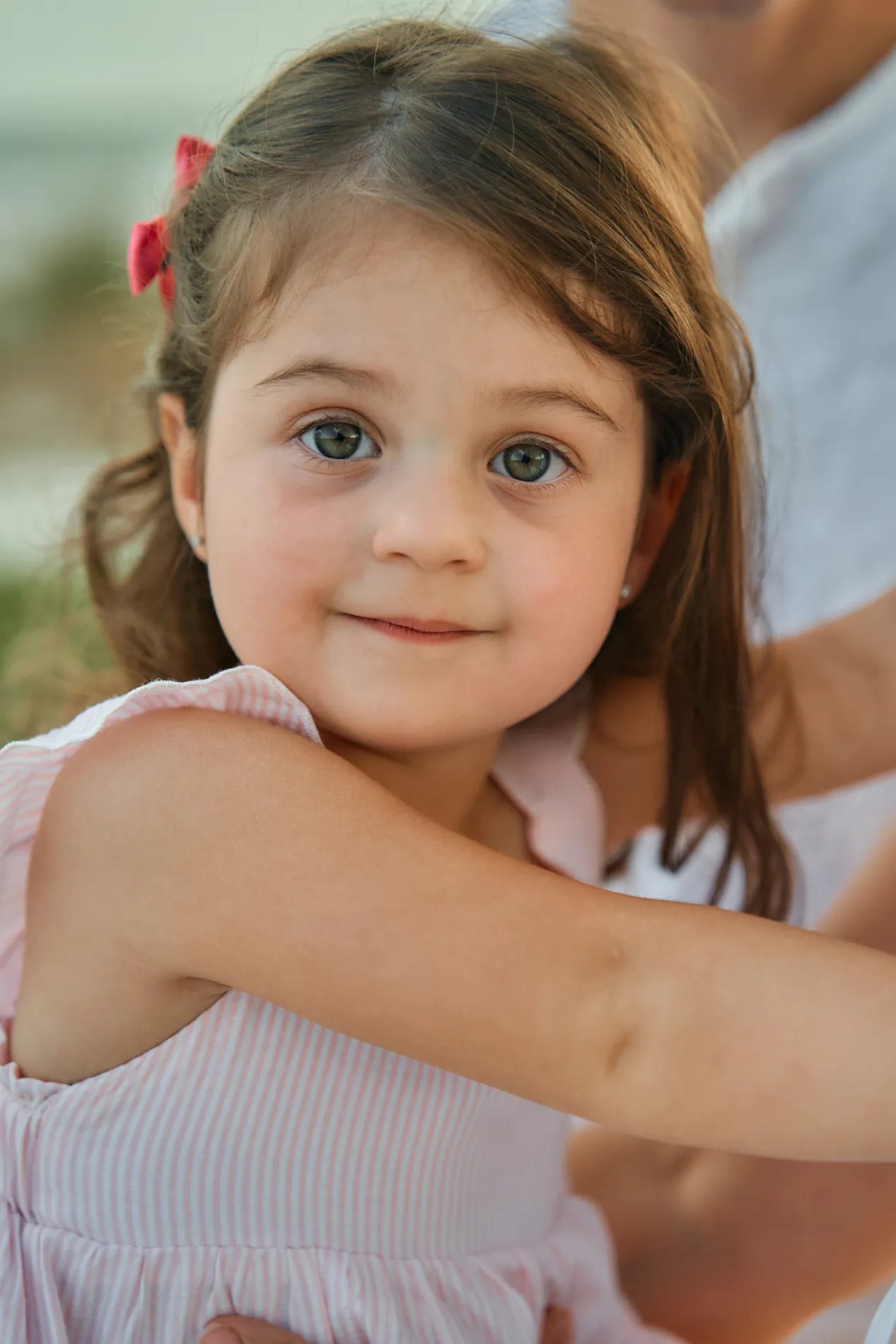 The same little girl close up, a pink hair clip and big grey eyes, looking straight at the camera while her father holds her