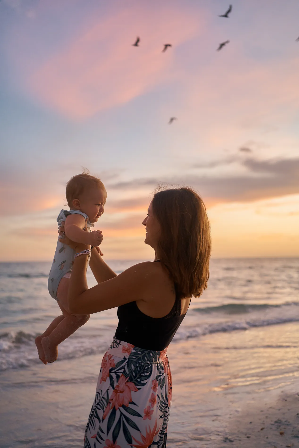A woman lifting a laughing baby above her head at the edge of the water on Siesta Key, pink and lilac clouds after sunset behind them