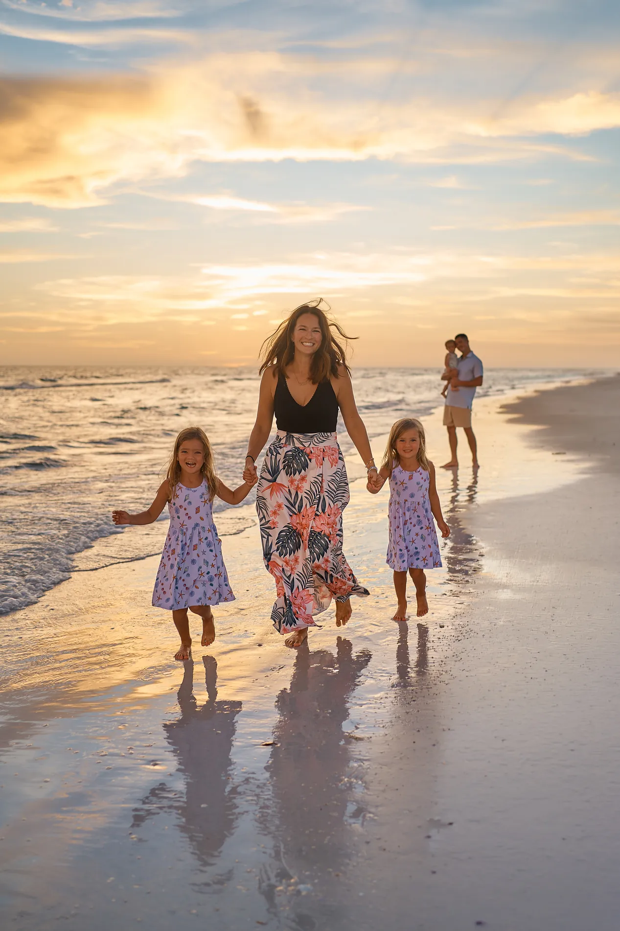 A mother in a flowered skirt walking two little girls in dresses by the hand along the water on Siesta Key, broken clouds lit gold and orange above them and their reflections on the wet sand