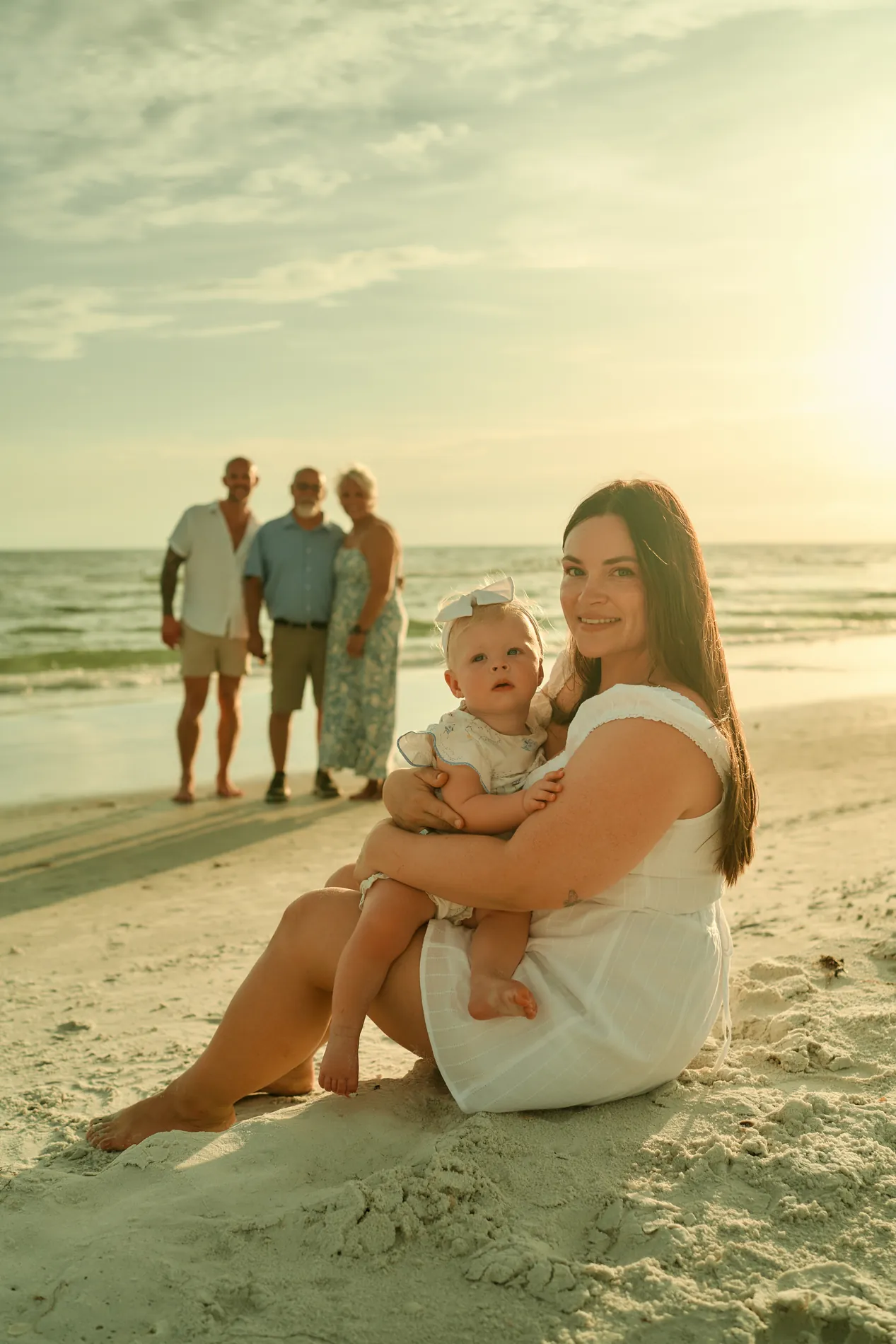 A young mother sitting on the sand with her baby on Siesta Key, the grandparents standing together at the water behind them in the low evening sun