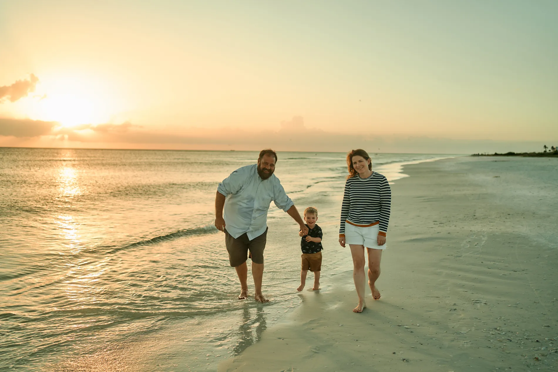 A man and a woman walking a toddler by both hands through the shallow water on Siesta Key, the low sun behind them over the Gulf