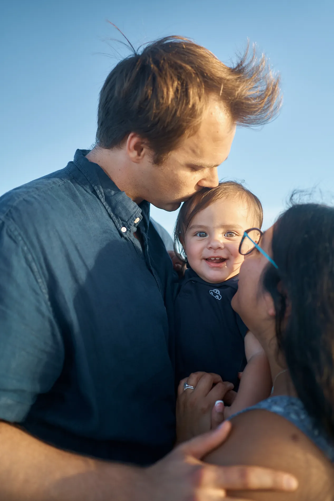A father with windblown hair kissing his little son on the head while the mother leans in beside them on Siesta Key