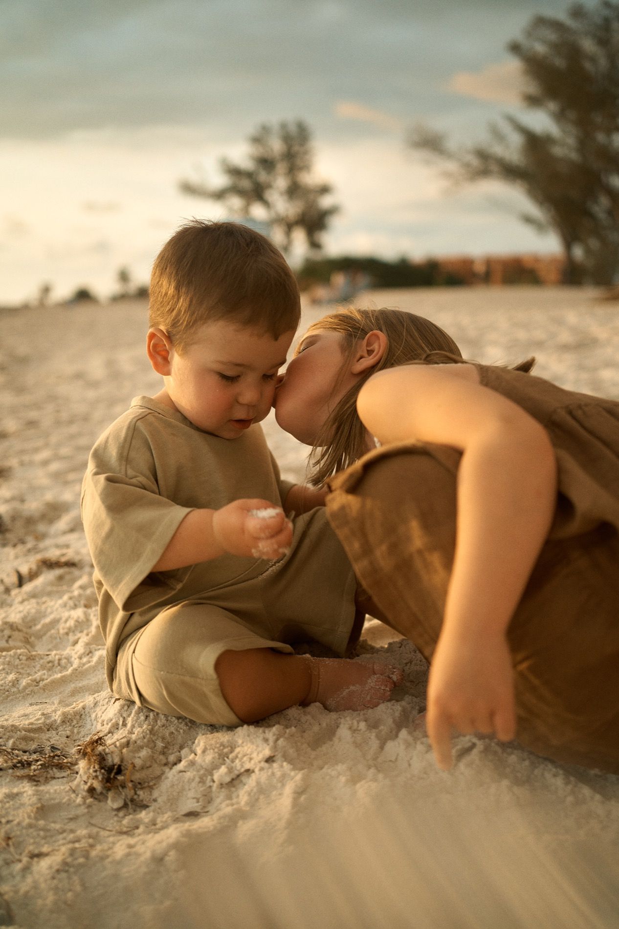 A big sister kissing her little brother on the sand at sunset